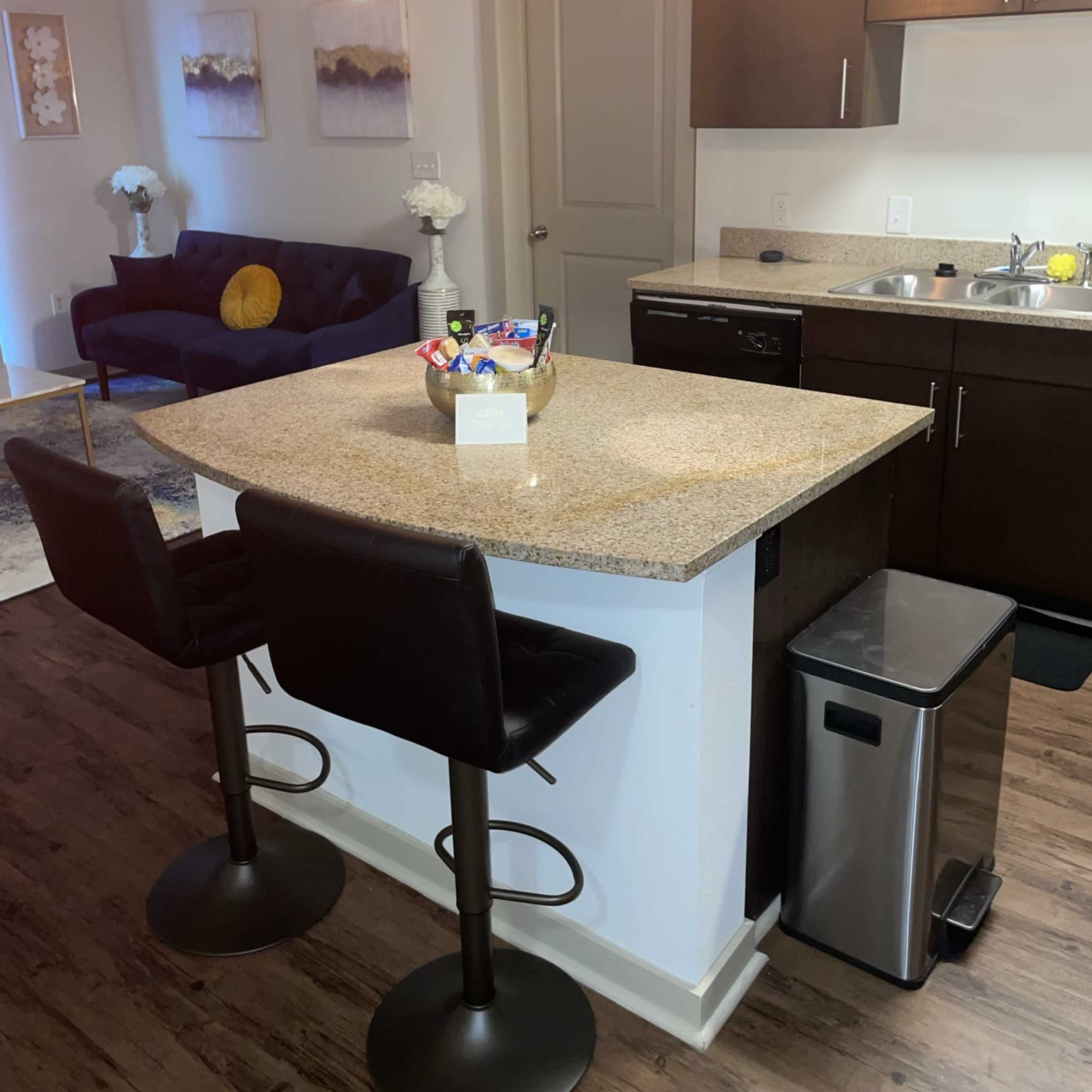 A modern kitchen with a granite island, two bar stools, and a small living area featuring a couch and decorative items.