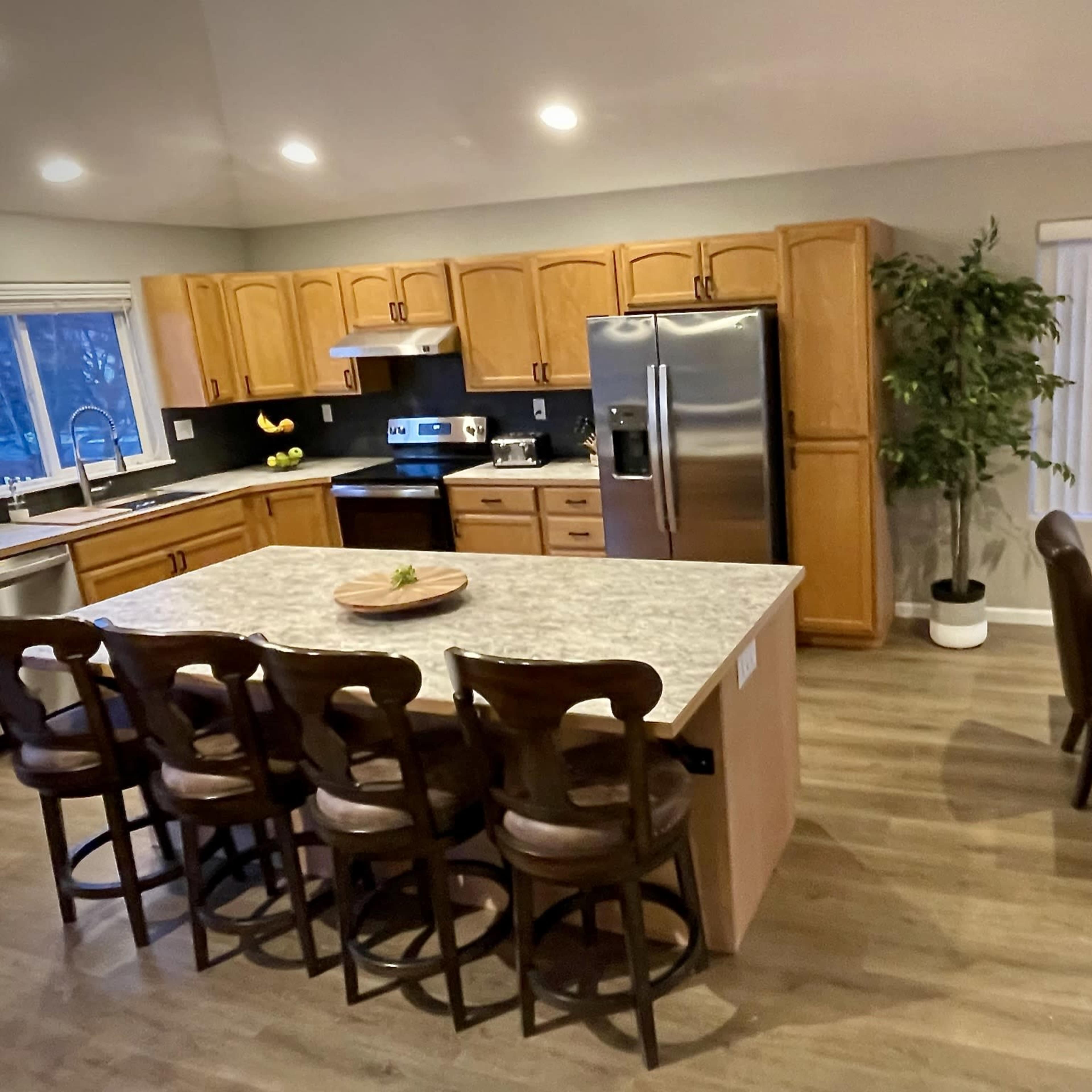 A modern kitchen with wooden cabinets, stainless steel appliances, a large island with bar stools, and a dining area featuring a wooden table and chairs.