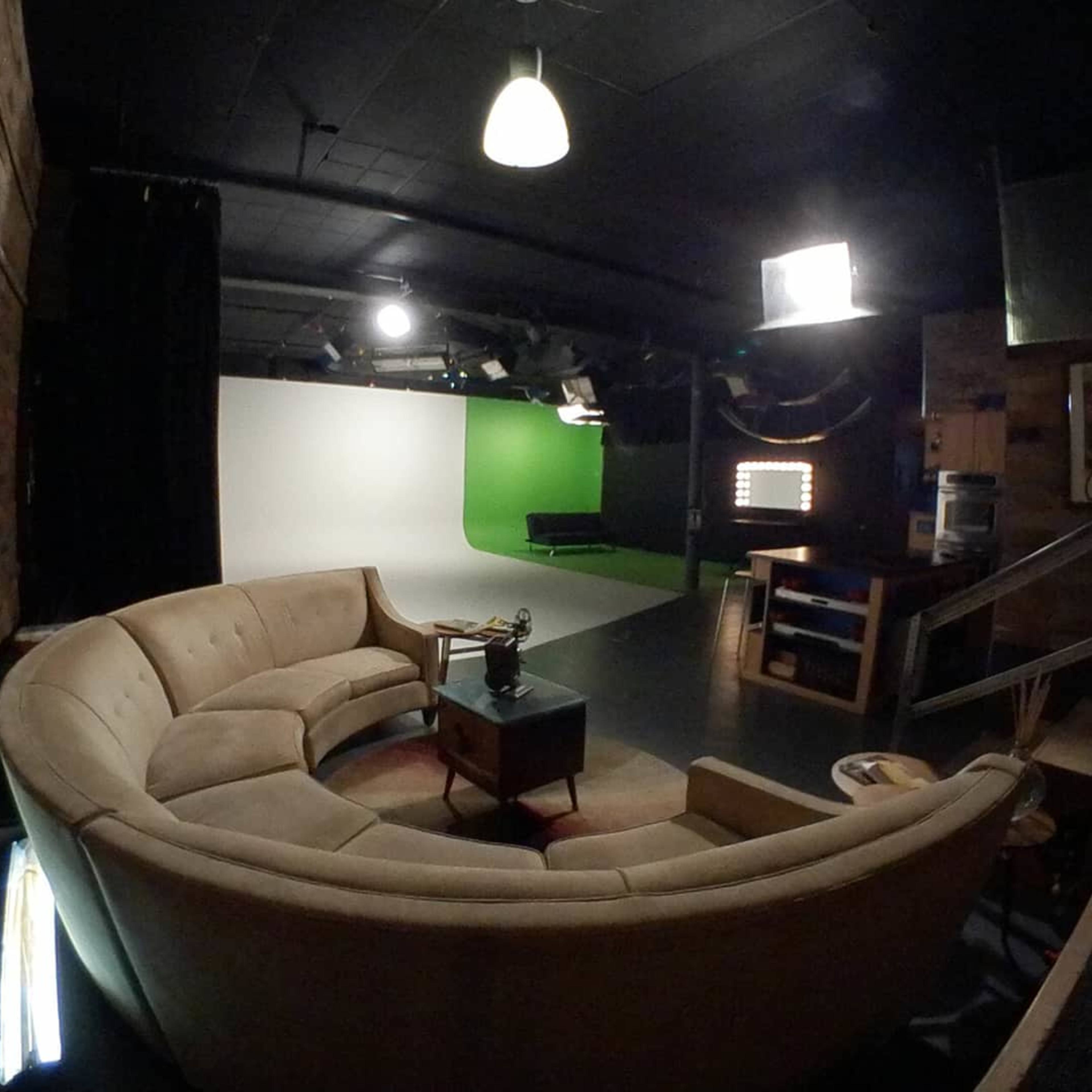 A studio space featuring a curved beige couch, a coffee table, and a backdrop with green screen and light fixtures.