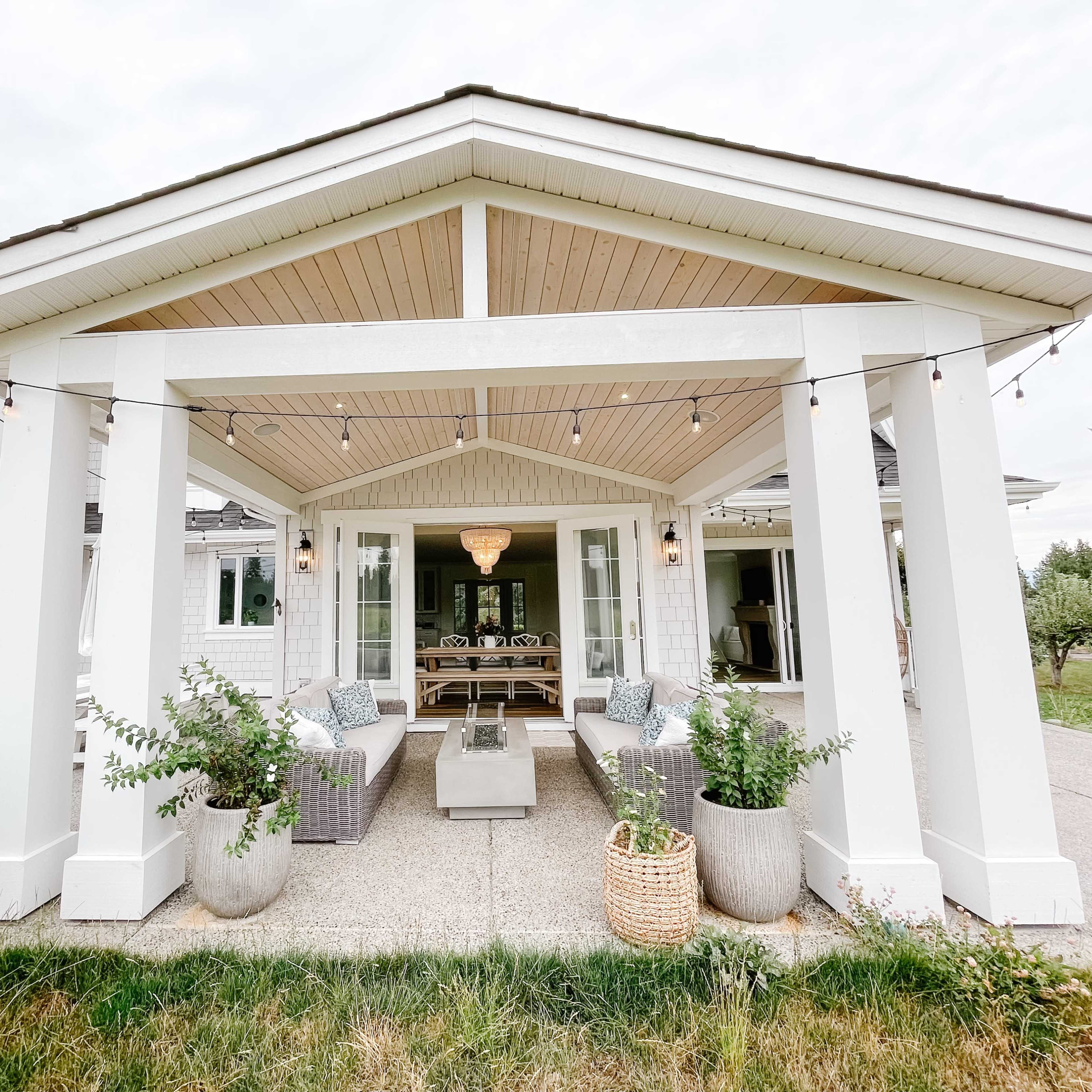 A covered outdoor patio with large white columns, seating arrangements, and potted plants, leading to a house in the background.