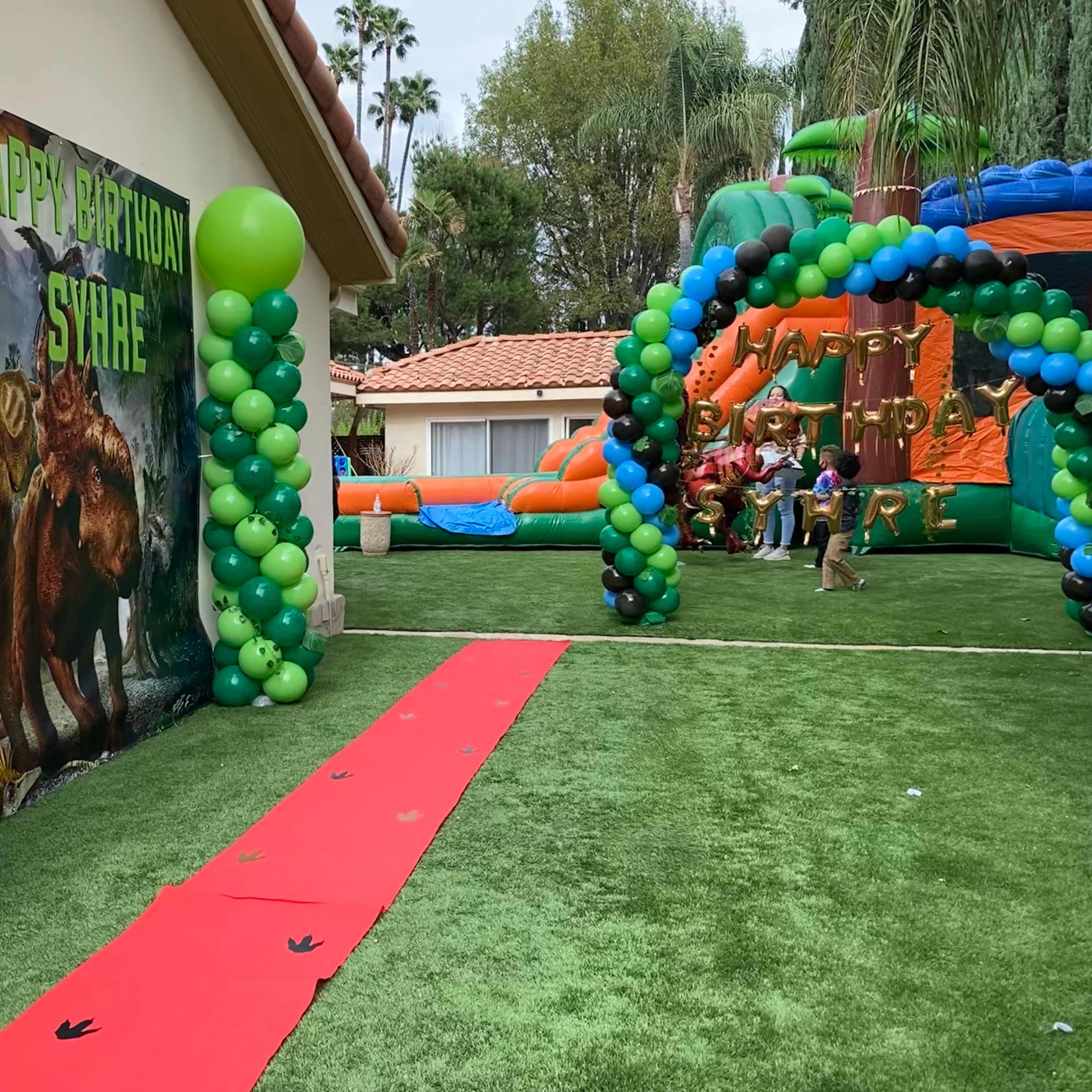 The scene features a decorated backyard for a birthday party, with green and blue balloon arches, a red carpet lined with dinosaur footprints, and a large dinosaur-themed backdrop.