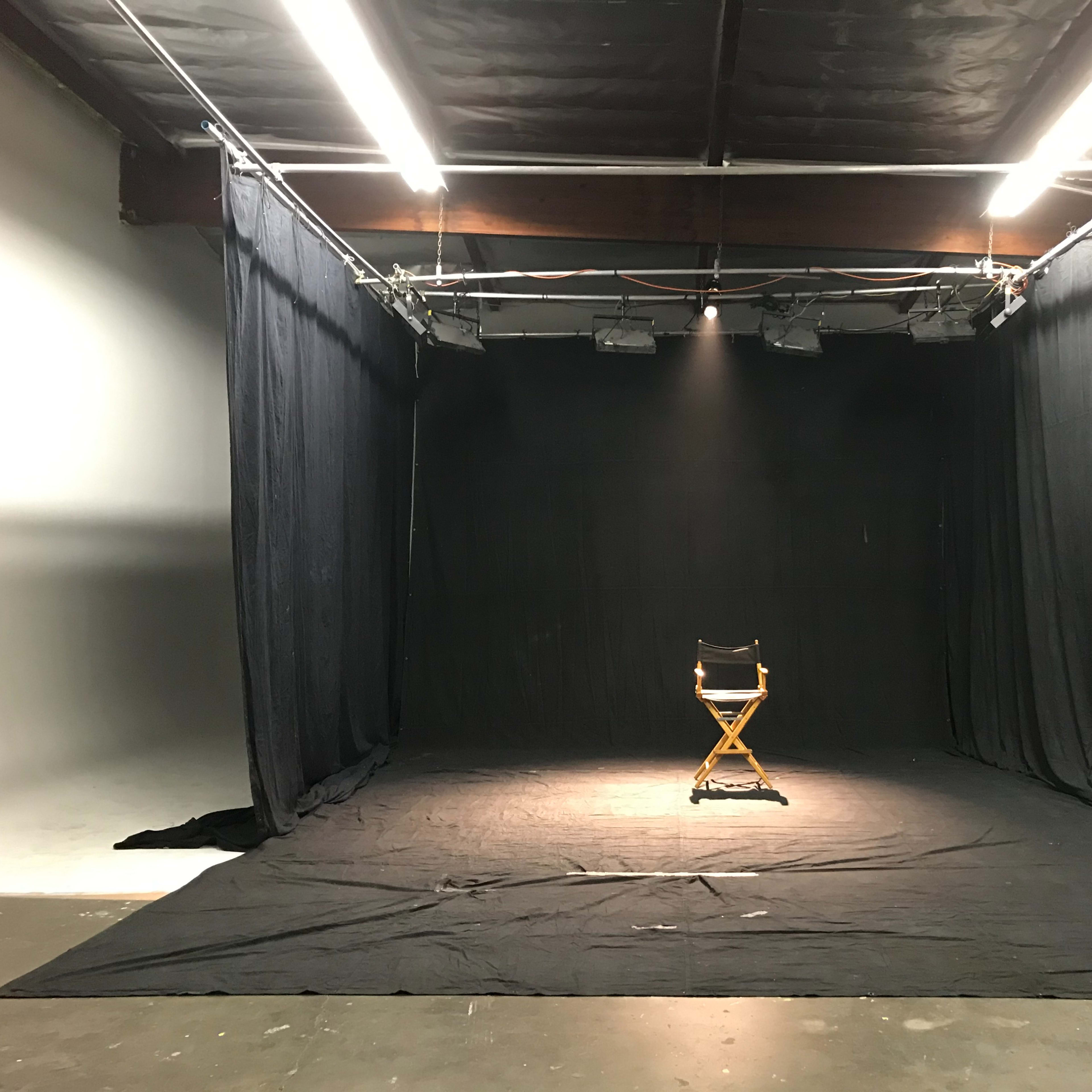 A spotlight illuminates an empty director's chair in a dimly lit studio surrounded by black curtains.