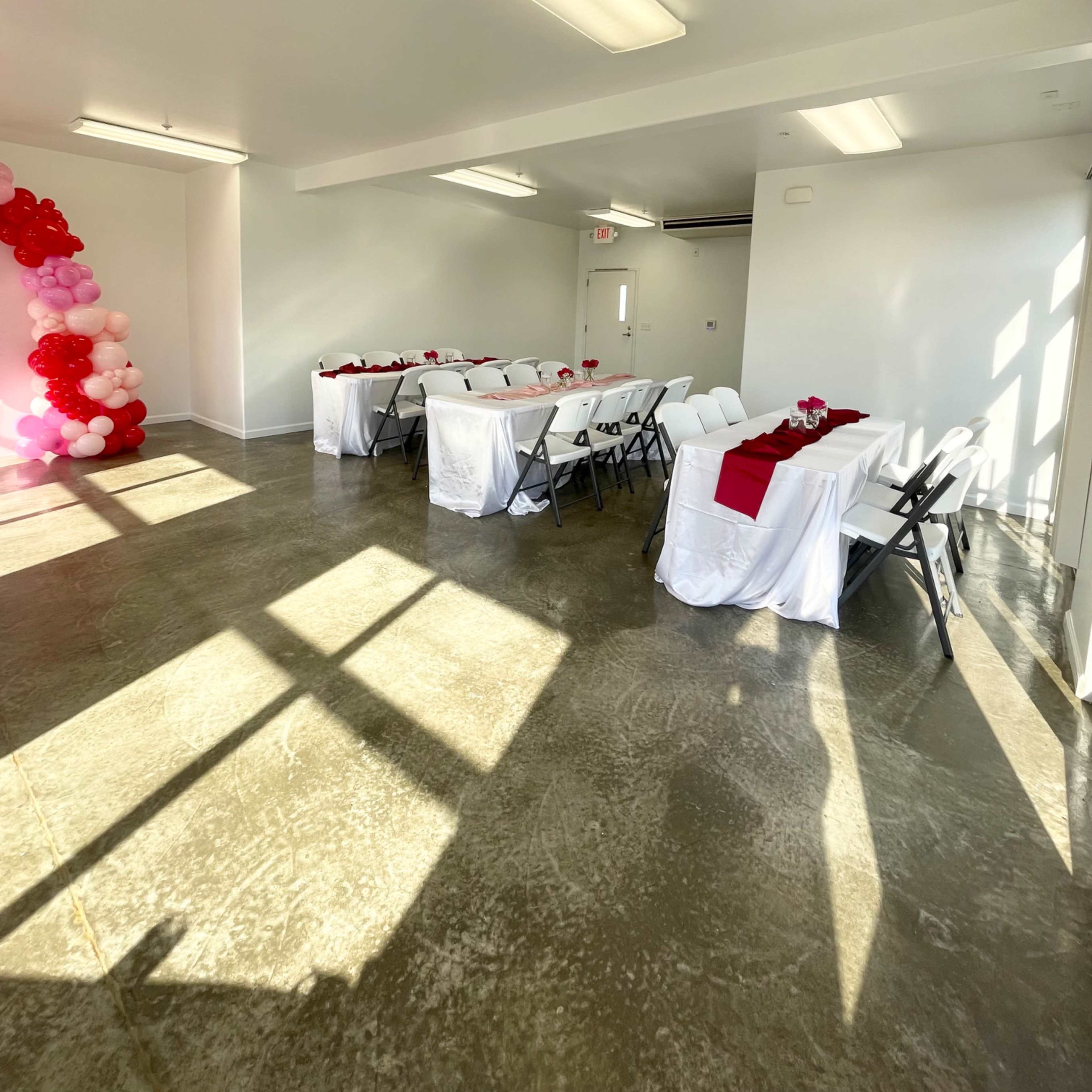 A room with two tables set for an event, each covered with white cloths and adorned with red accents, illuminated by natural light streaming through windows.
