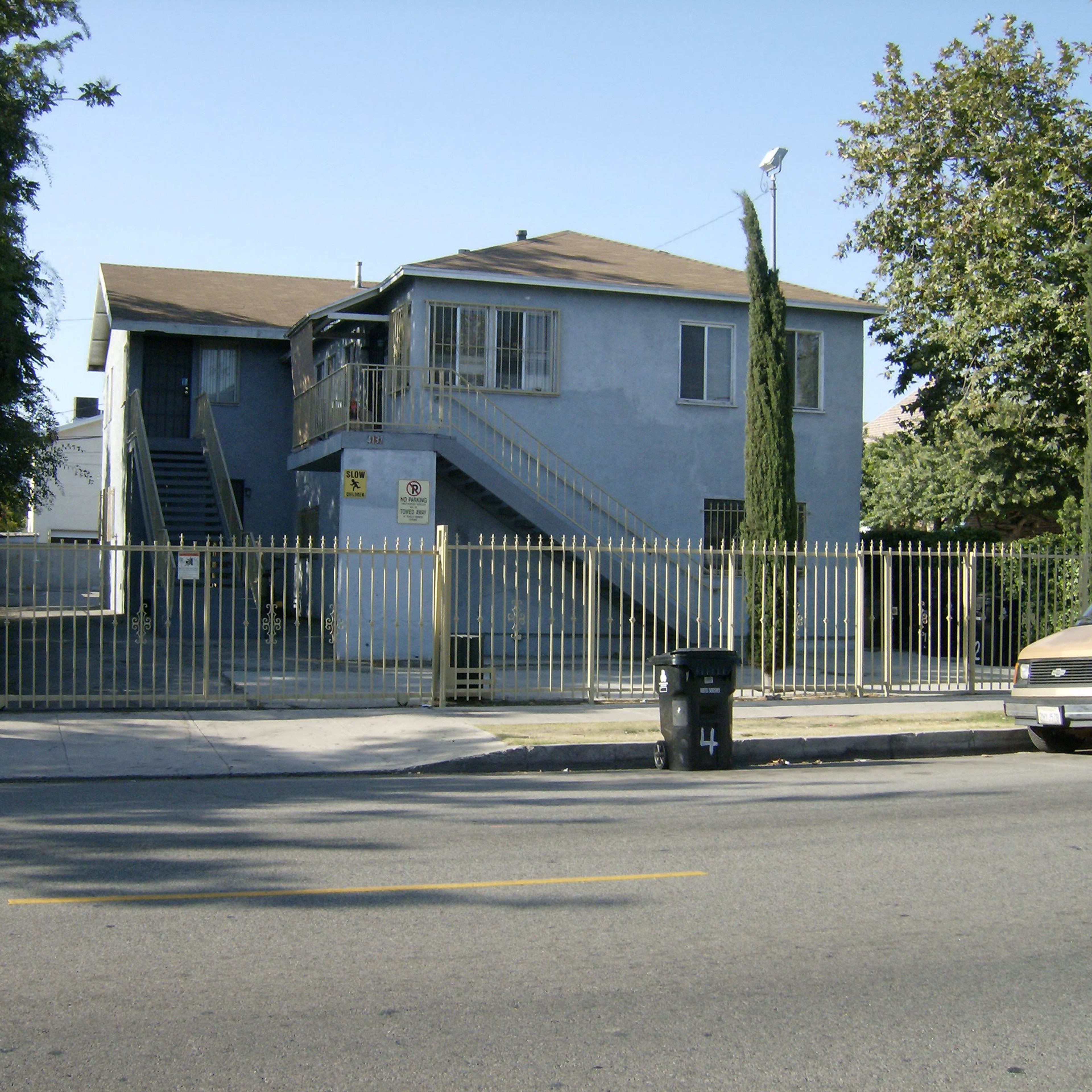 A two-story blue residential building with a fenced yard and a staircase on the side, located along a street.
