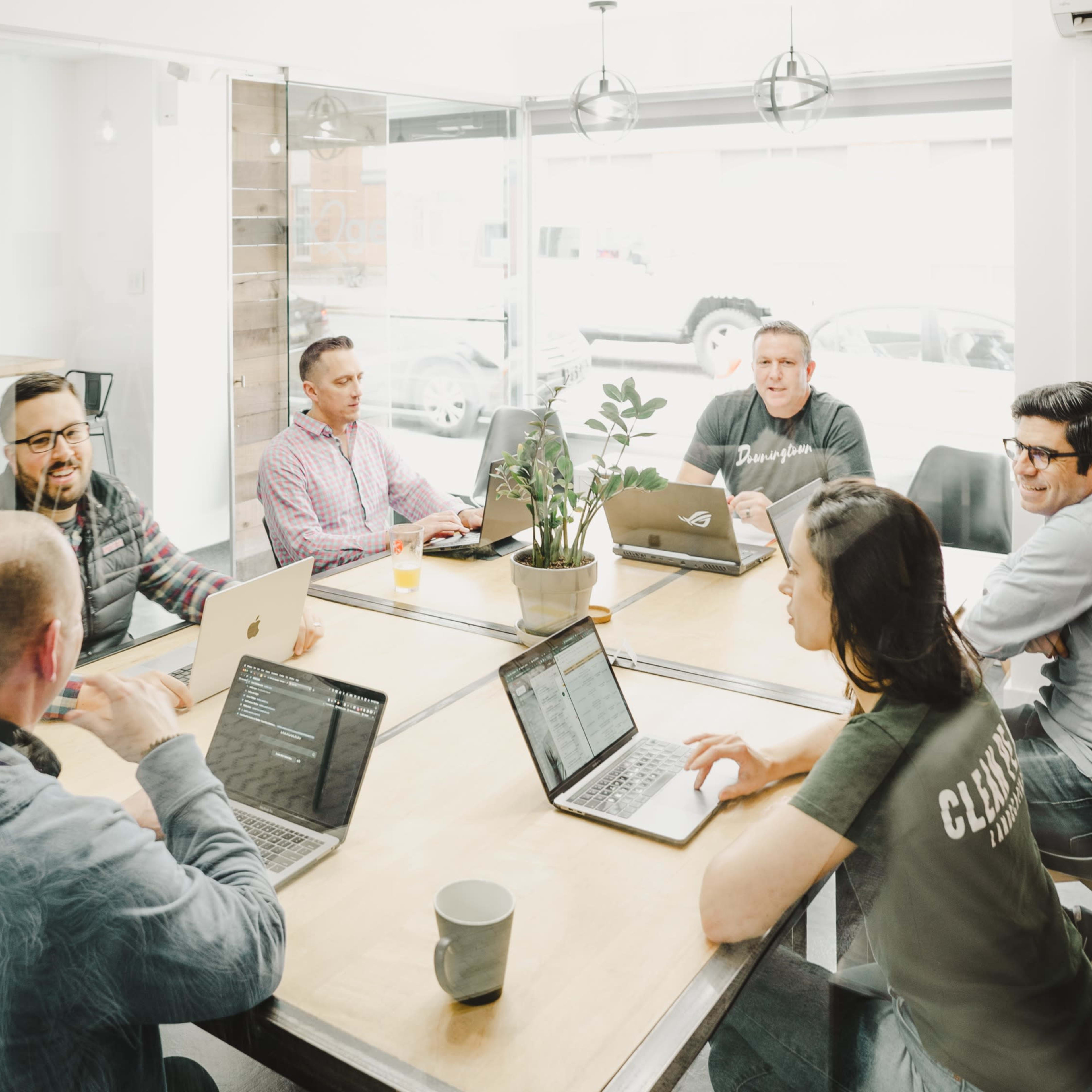 A group of six people is seated around a wooden table in a bright meeting room, working on laptops and discussing ideas.