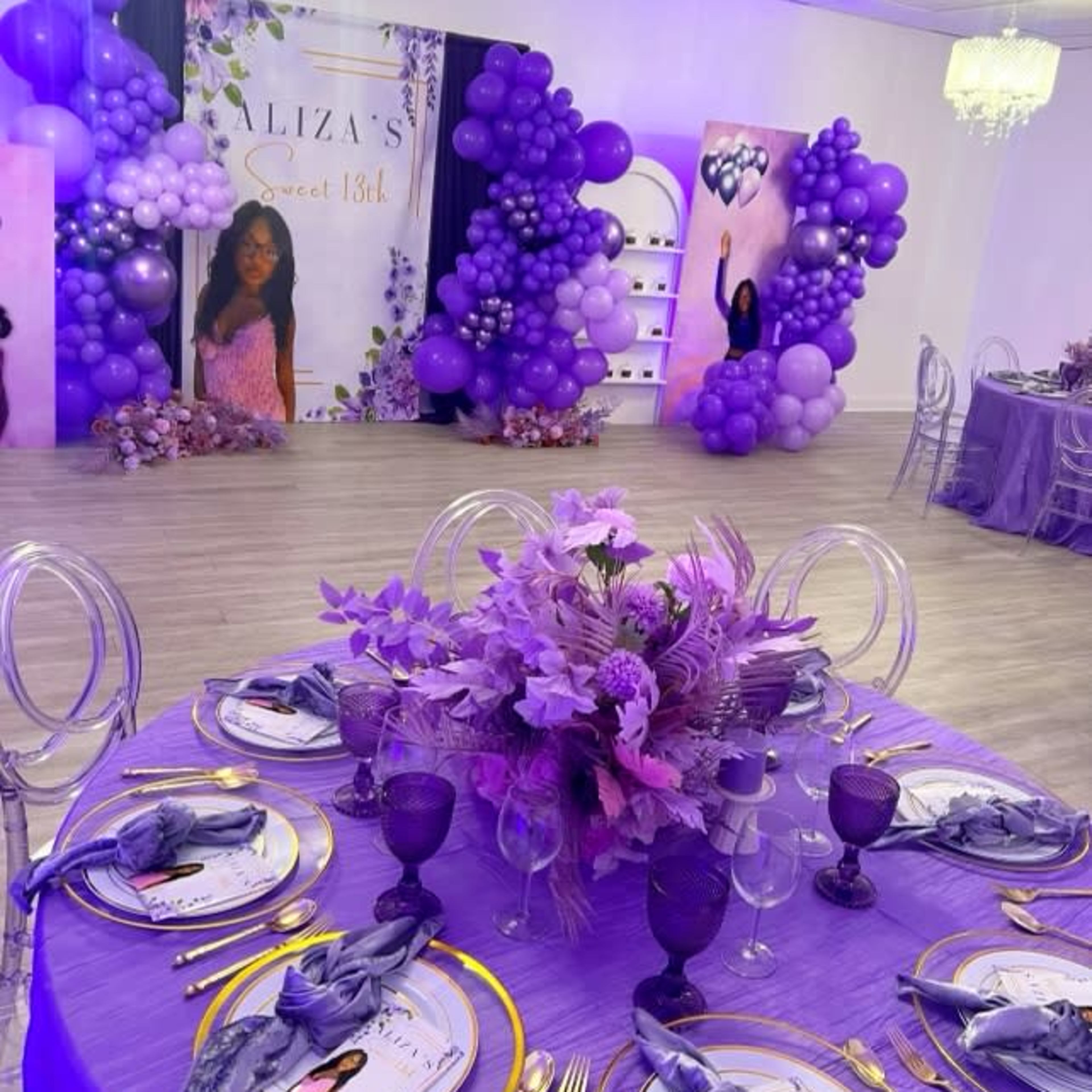 A decorated event space for a sweet 18th birthday celebration, featuring purple-themed decorations, table settings, and a backdrop with balloons and a portrait.