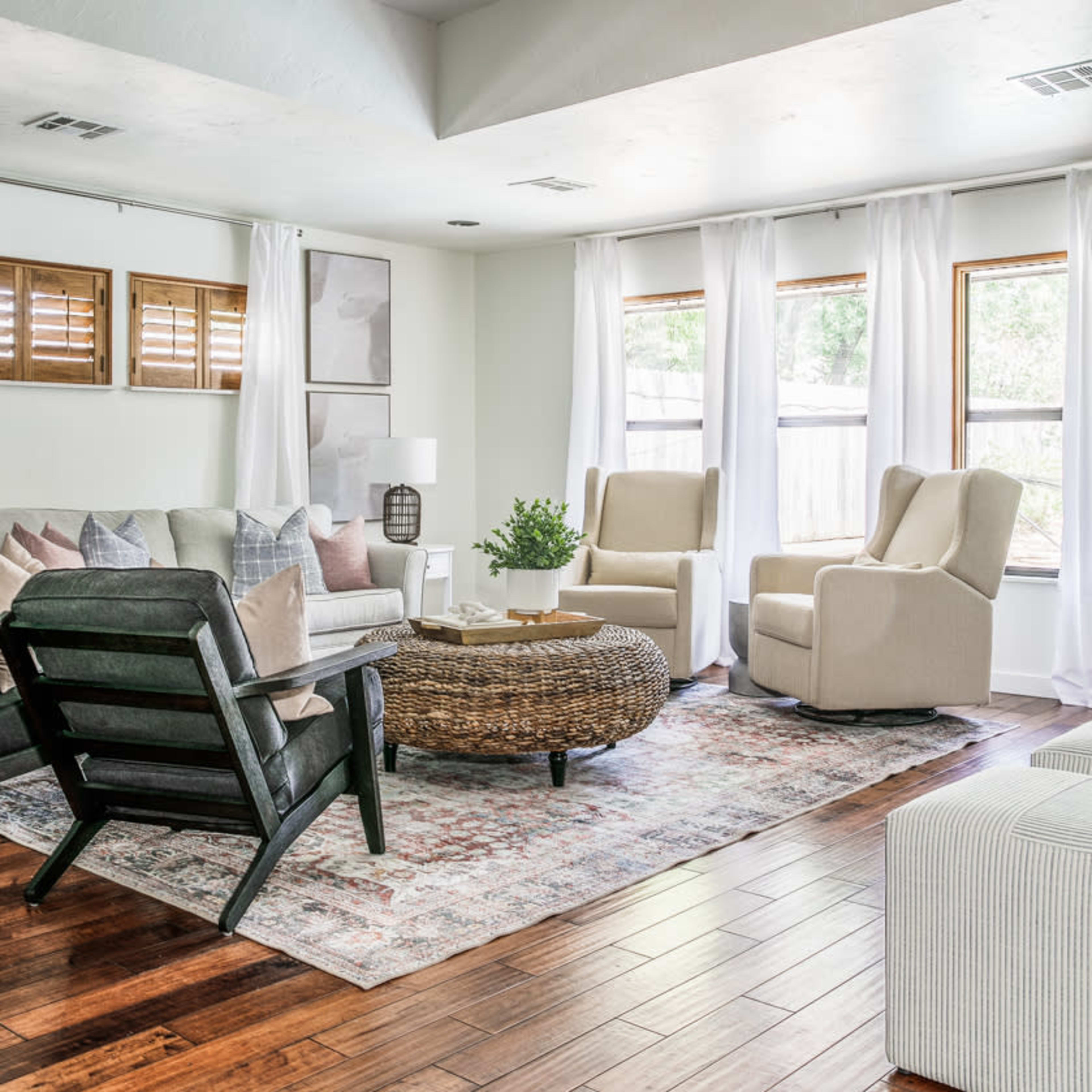 A living room features two armchairs, a round coffee table, a sofa, and large windows with sheer curtains, all set on a hardwood floor.