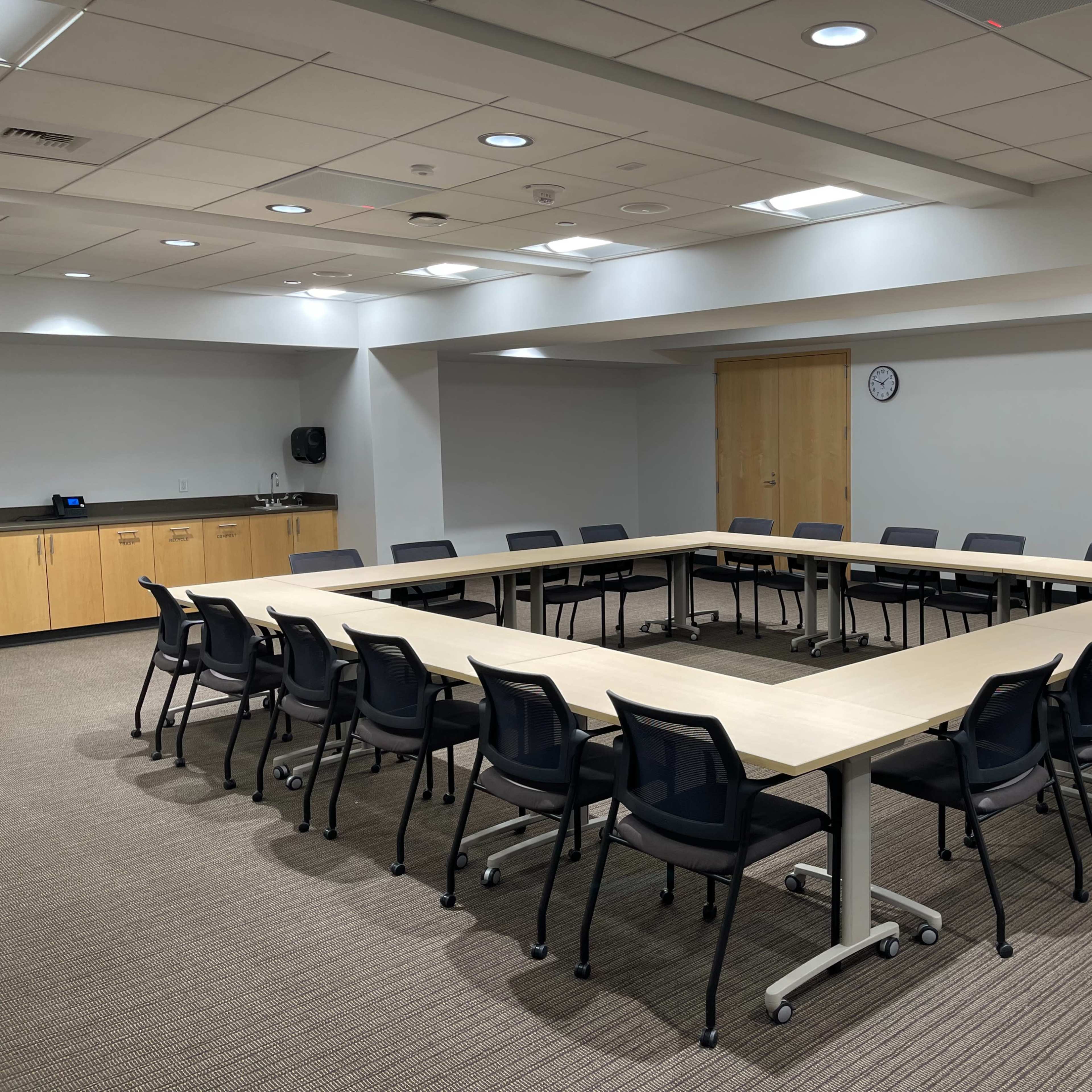 A large conference room features a long table arranged in an open square shape, surrounded by black chairs, with wooden cabinets and a clock visible on the walls.