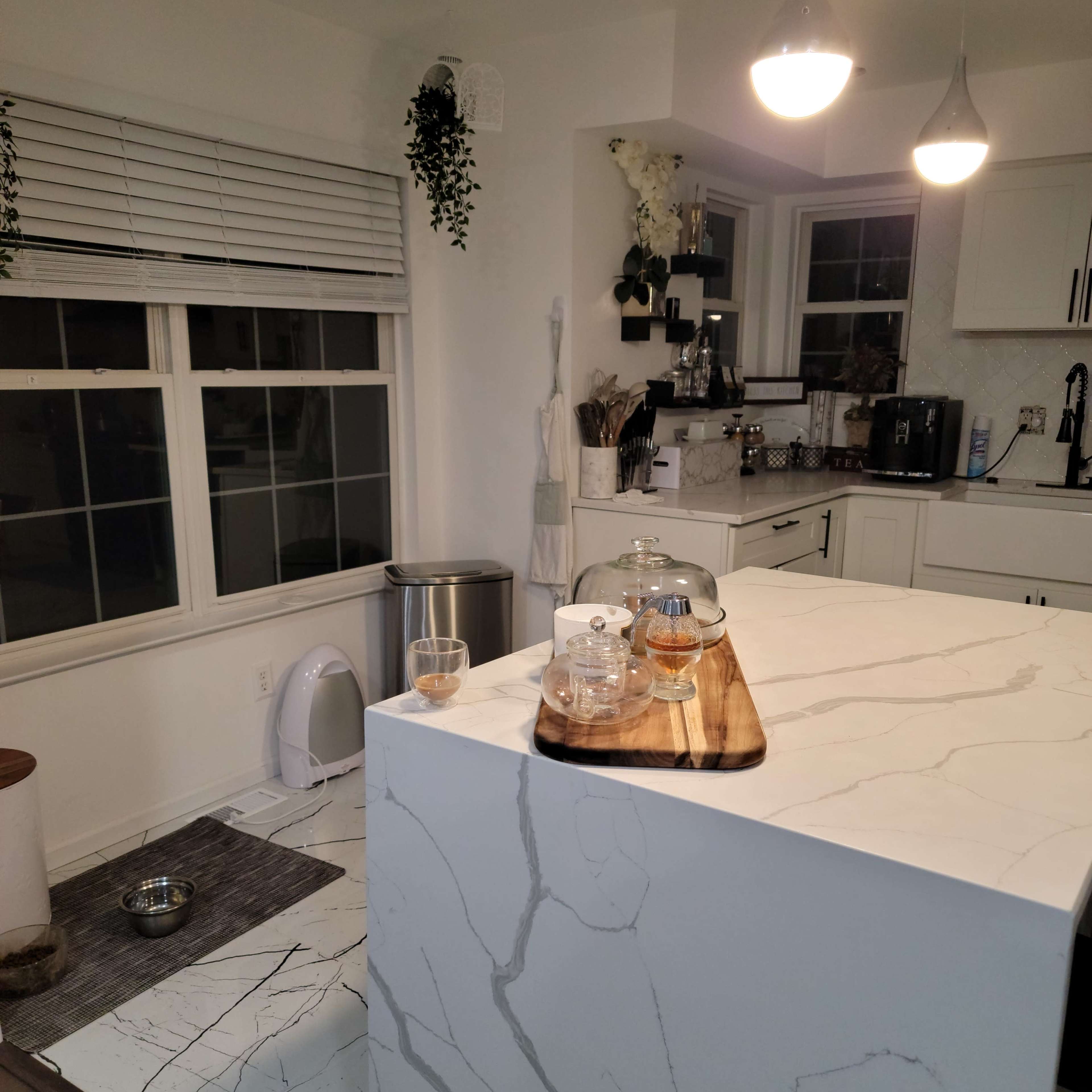 A modern kitchen with a marble island, a wooden serving tray with glass containers, and a window beside the sink, featuring a tidy layout and a pet's food bowl on the floor.