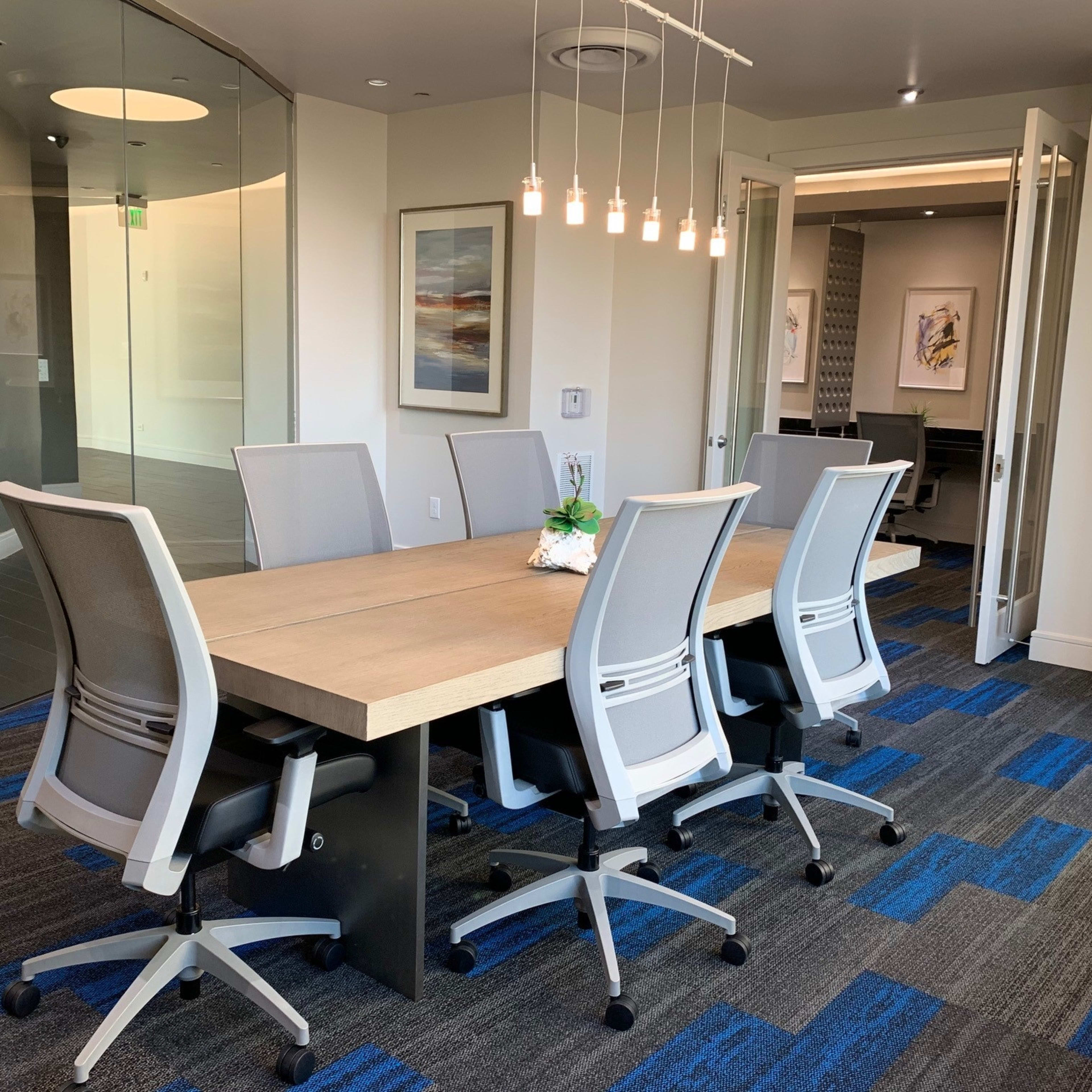 A modern conference room features a long table surrounded by six ergonomic chairs, with a glass wall and hanging light fixtures above.