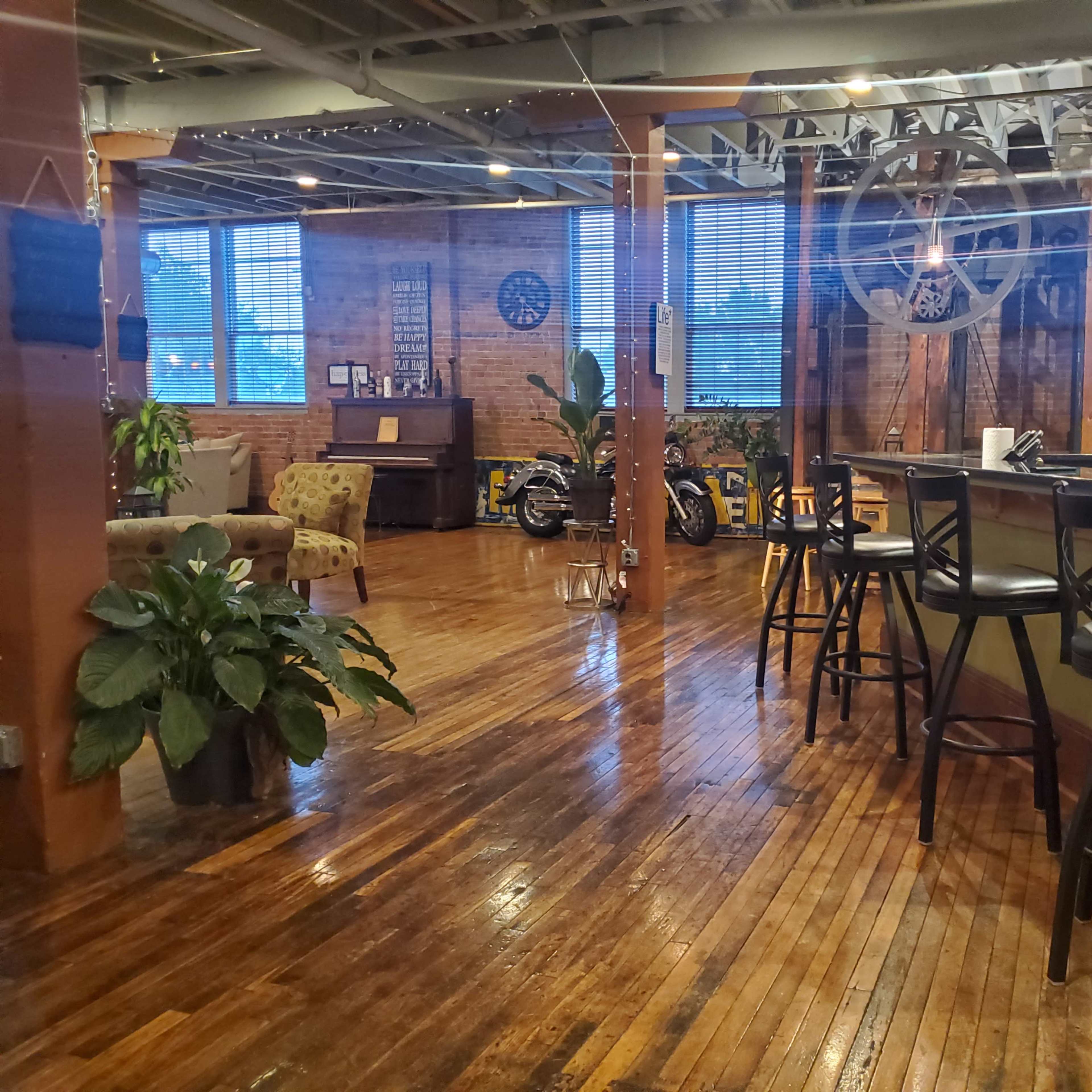 A spacious, industrial-style interior with wooden floors, a bar area, plants, and a decorative wall featuring a large wheel.