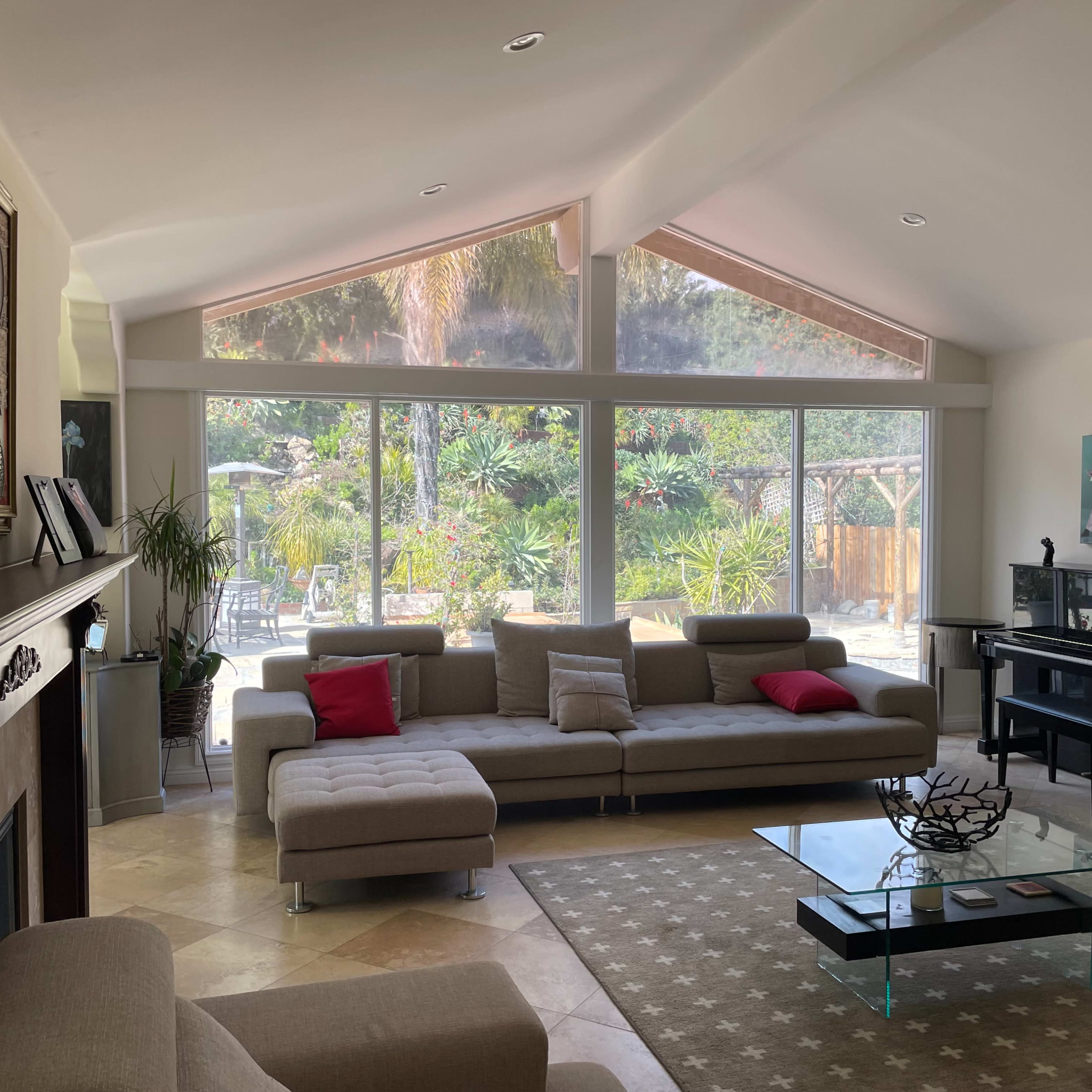 The living room features a large sectional sofa, a coffee table, and a grand piano, with expansive windows overlooking a garden area.