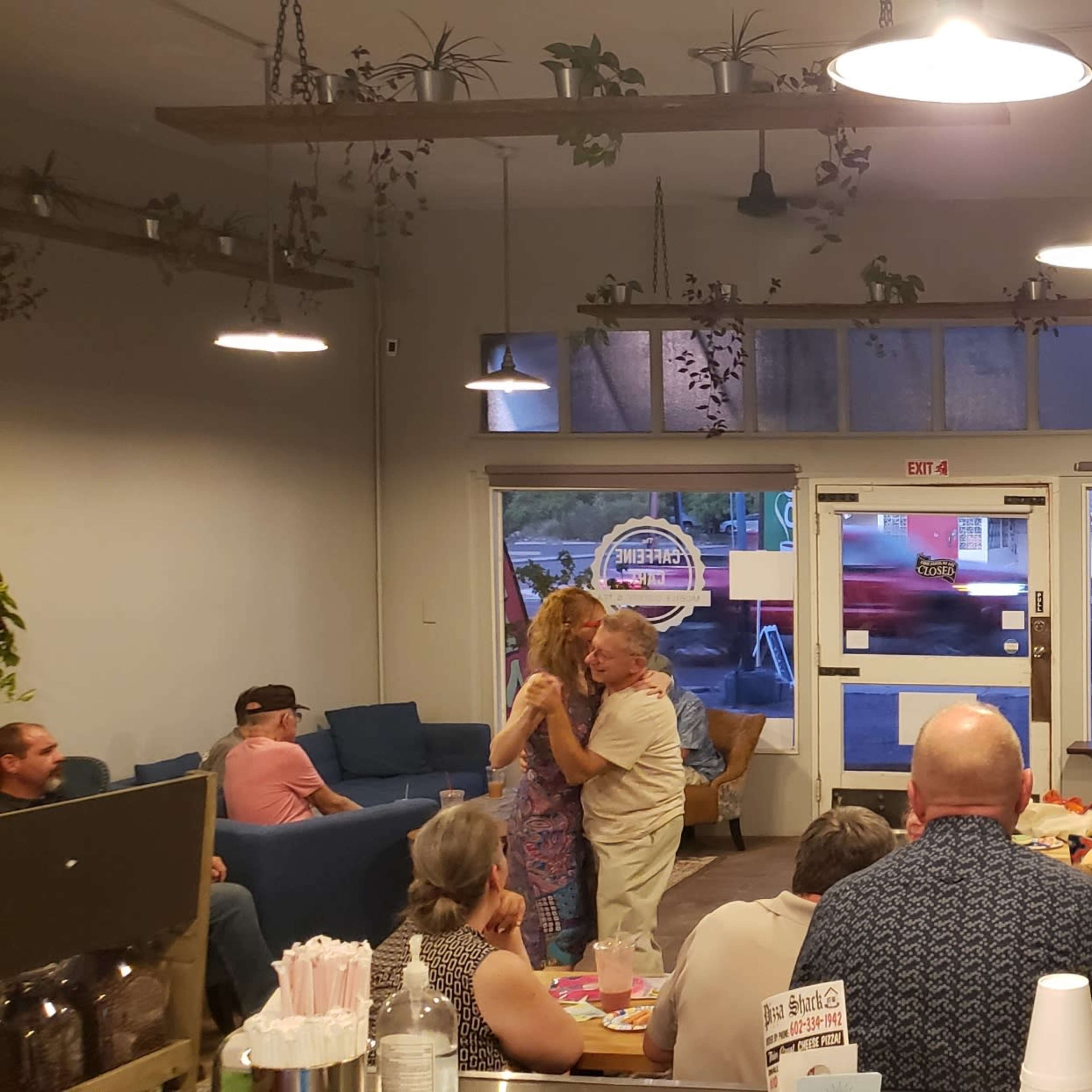 A group of people gathers in a brightly lit café with colorful decor and a mural, engaging in conversation and dancing near a window displaying parked cars outside.