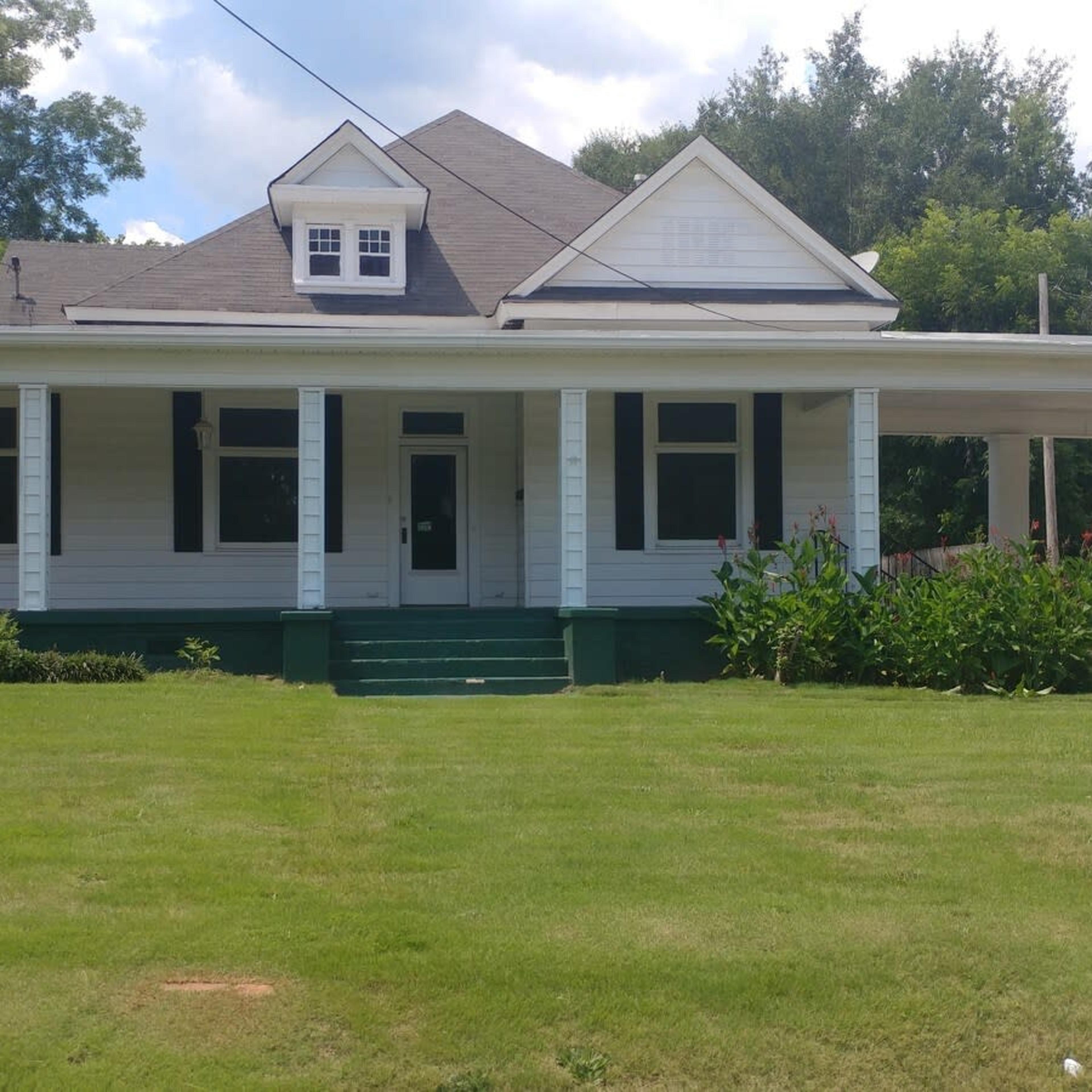 A two-story white house with a covered porch and green steps is surrounded by grass and shrubs.