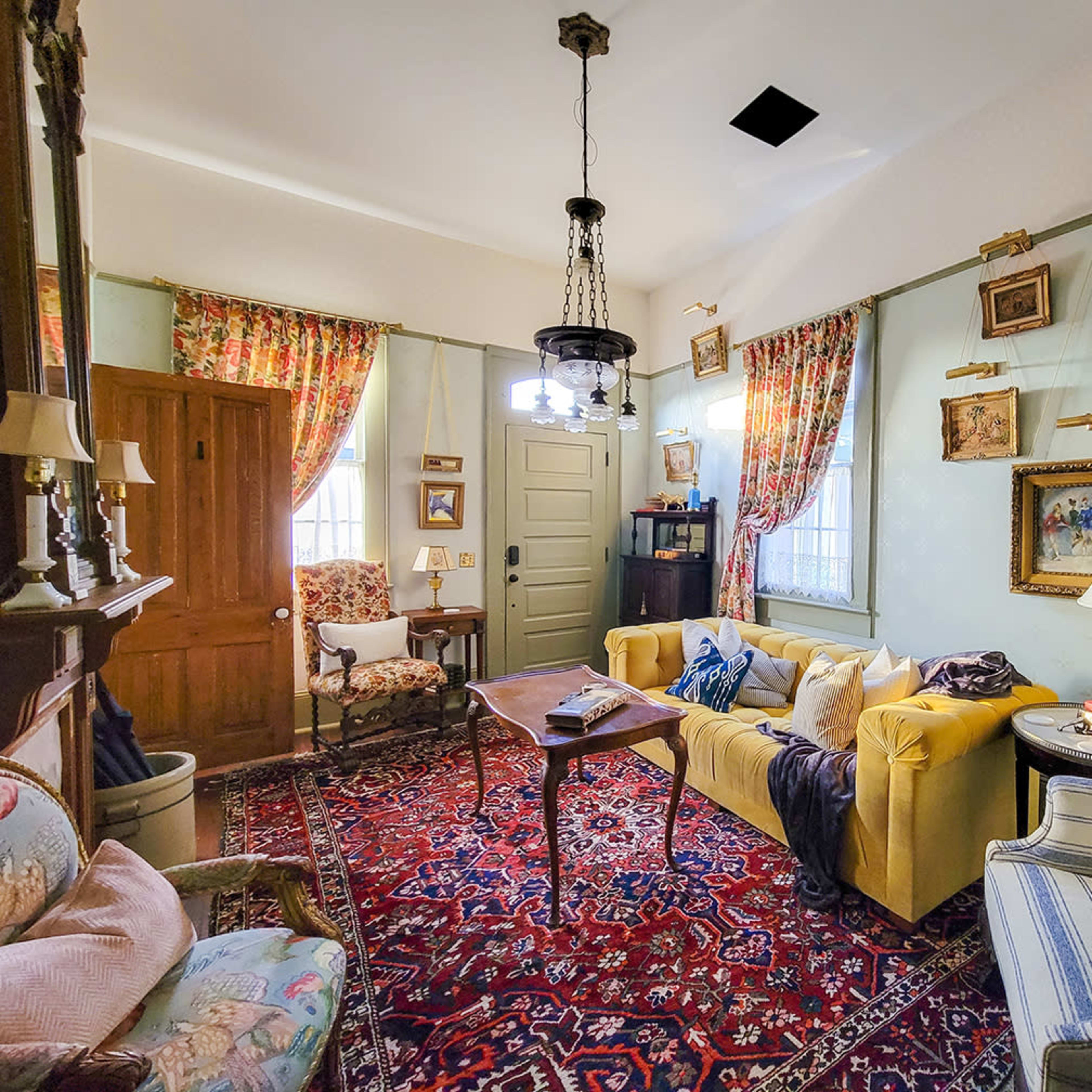 The room features a yellow sofa, two patterned armchairs, a wooden coffee table, and various pieces of artwork hanging on the walls, complemented by floral curtains and a large area rug.