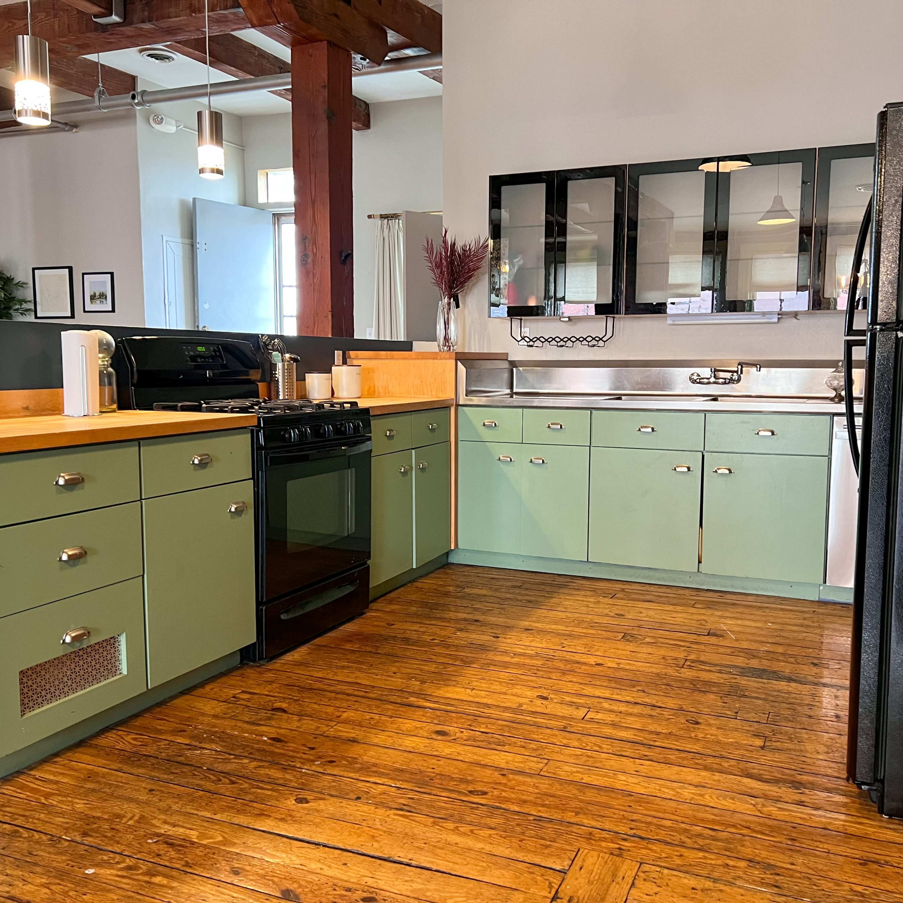 A kitchen features green cabinetry, a black stove, a stainless steel sink, and a black refrigerator, with wooden floors and overhead lighting.