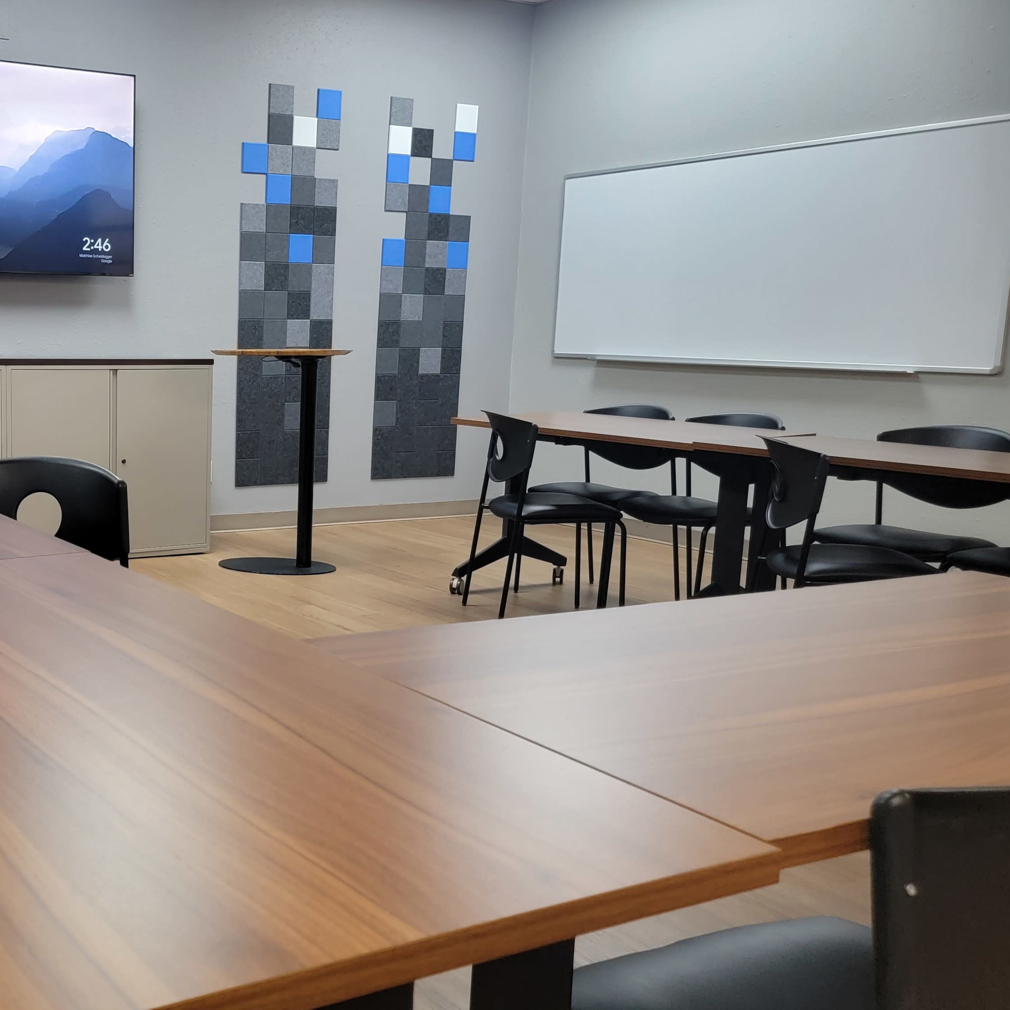 A modern conference room with a long wooden table, several black chairs, a wall-mounted television, and a whiteboard.