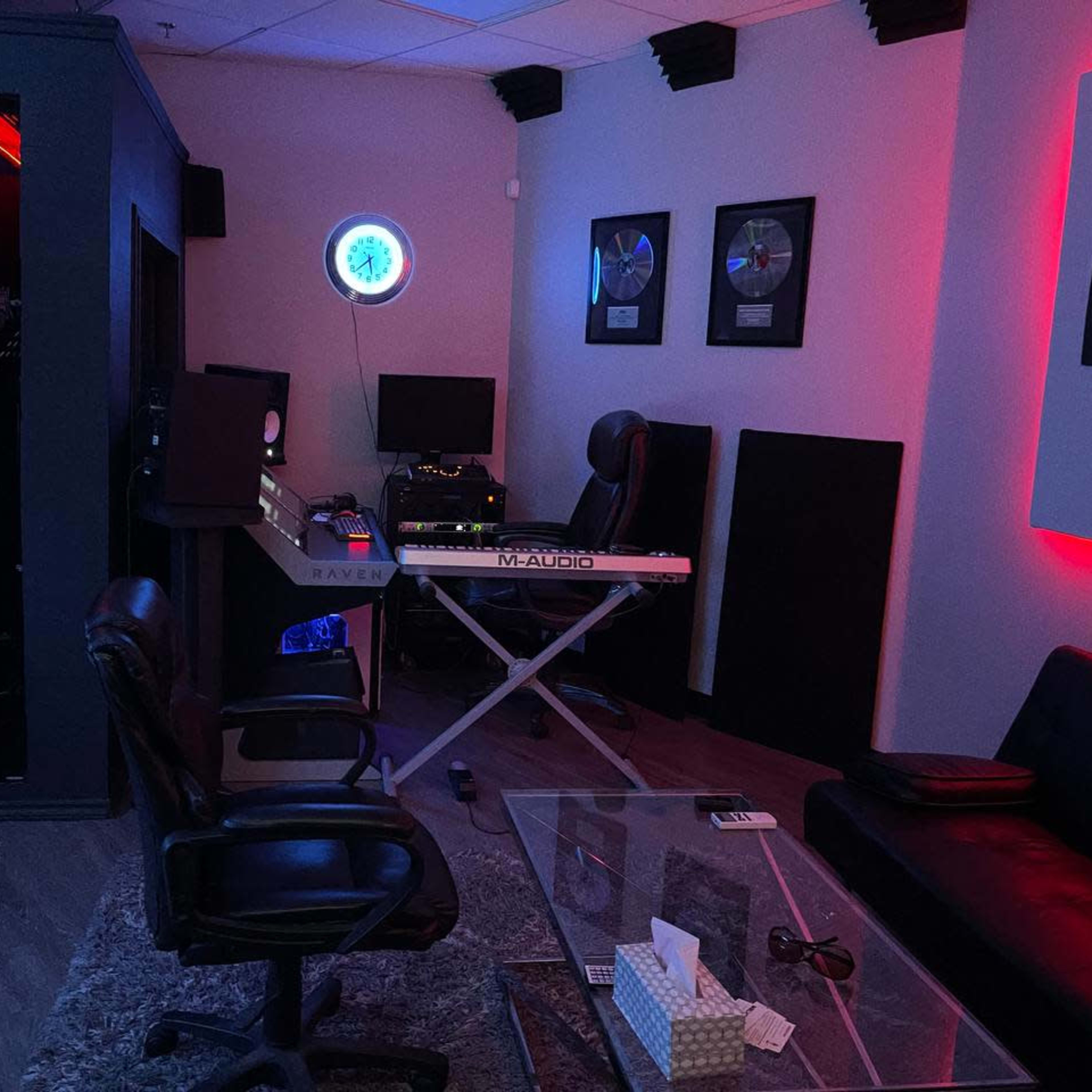 A music studio with a mixing desk, keyboard, and soundproof booth, illuminated by red and blue lighting.