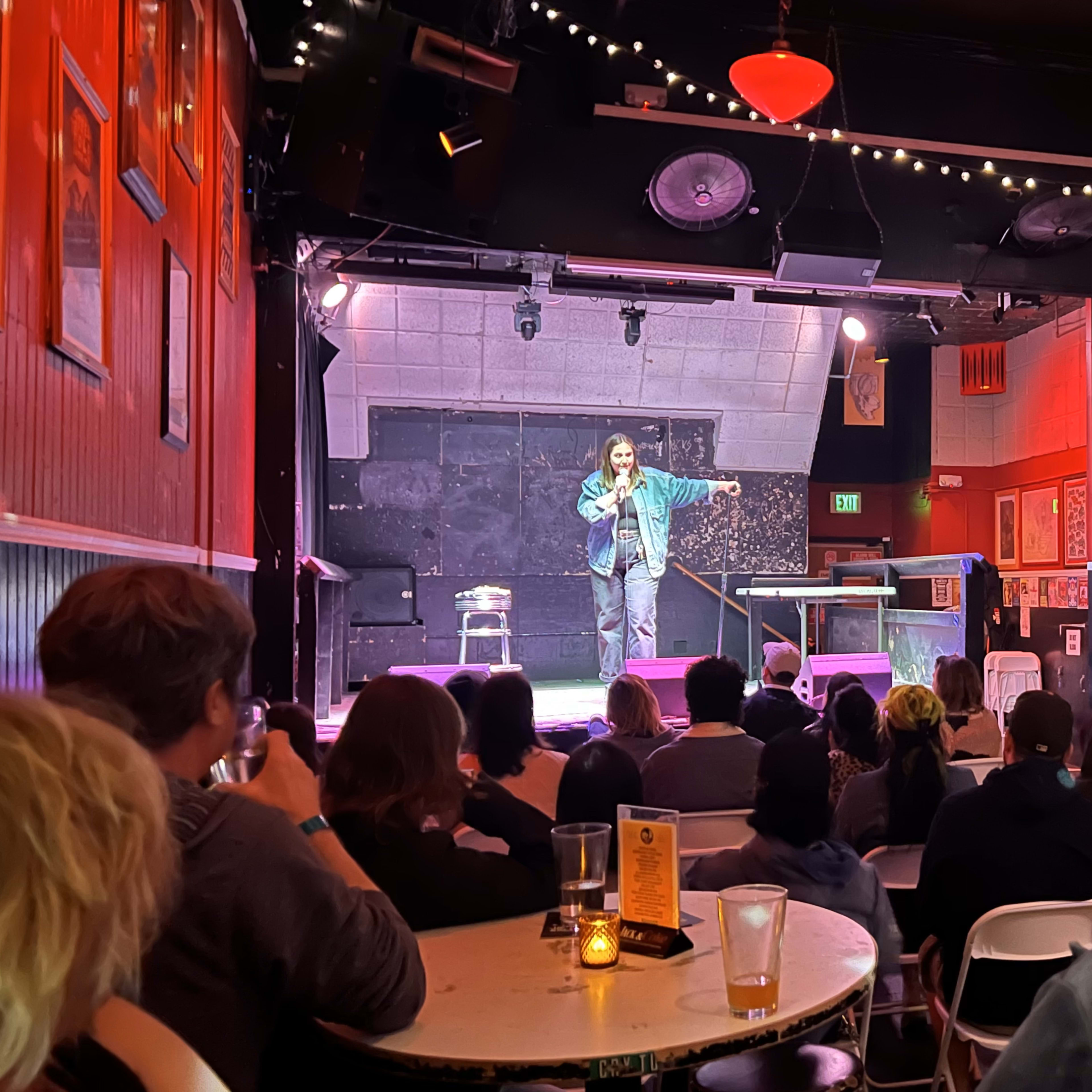 A comedian performs on stage in a dimly lit comedy club while an audience watches from tables.