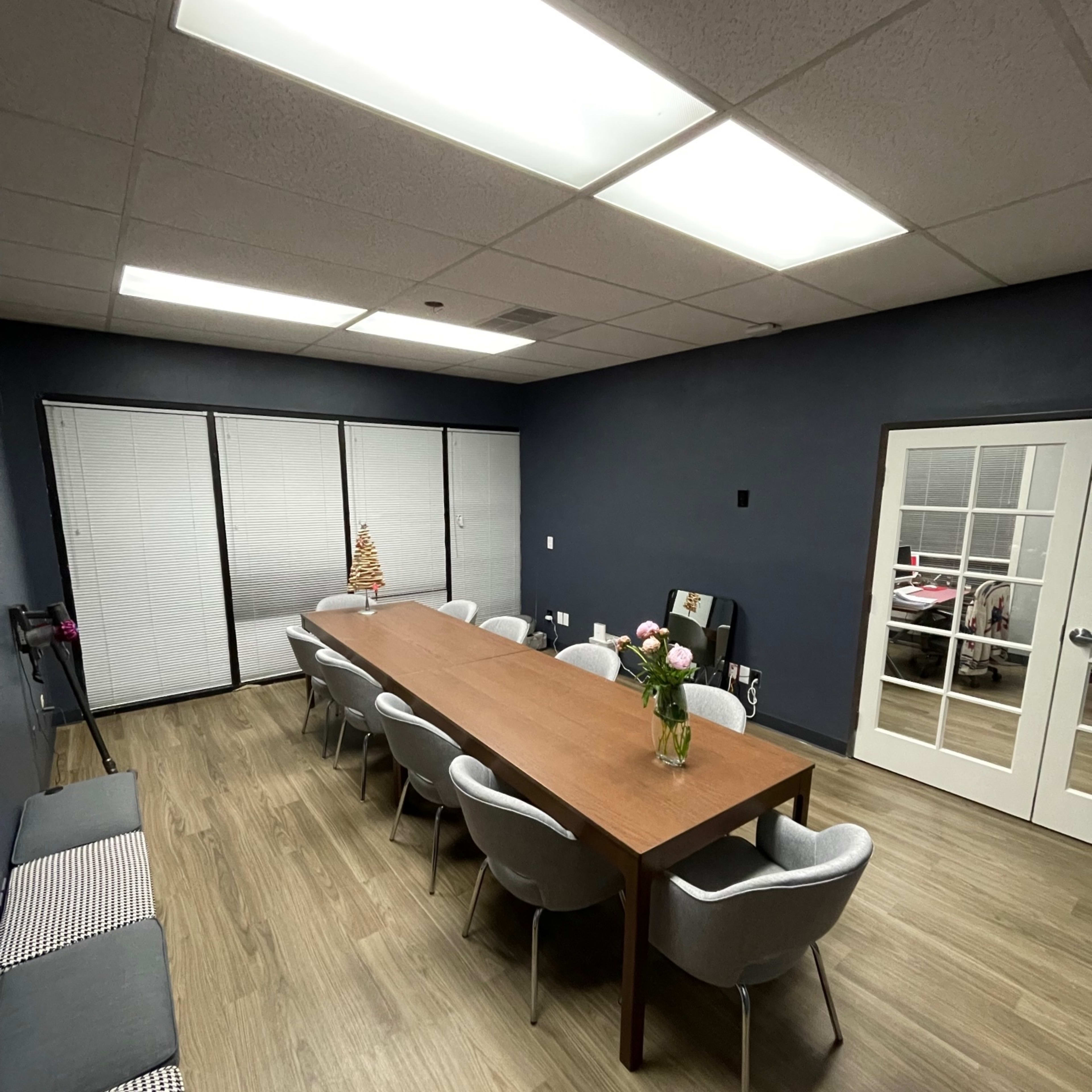 A long wooden table is surrounded by chairs in a well-lit conference room with dark blue walls and large windows covered by blinds.