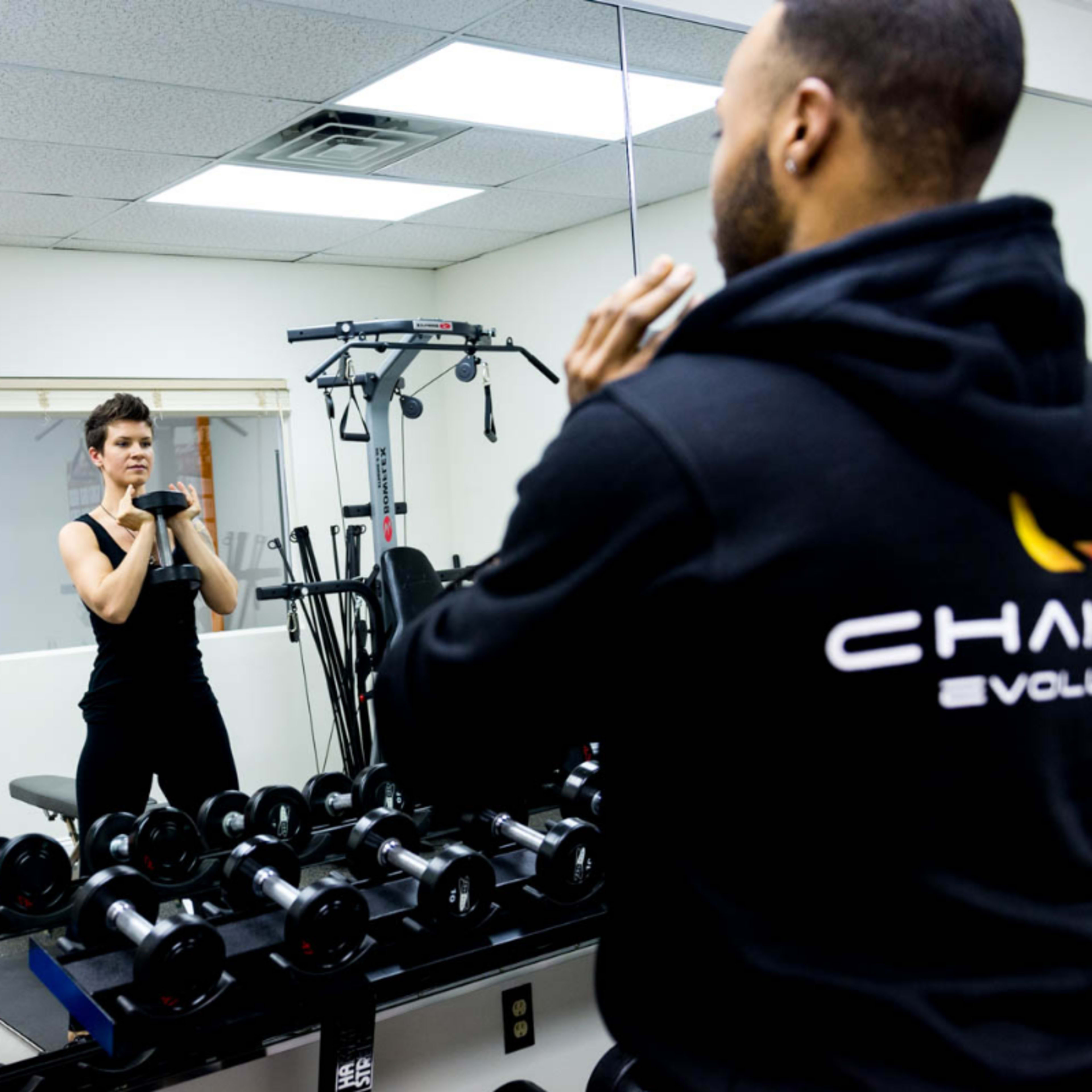 Two people are performing a weightlifting exercise with dumbbells in a gym, while a third person observes them in the mirror.