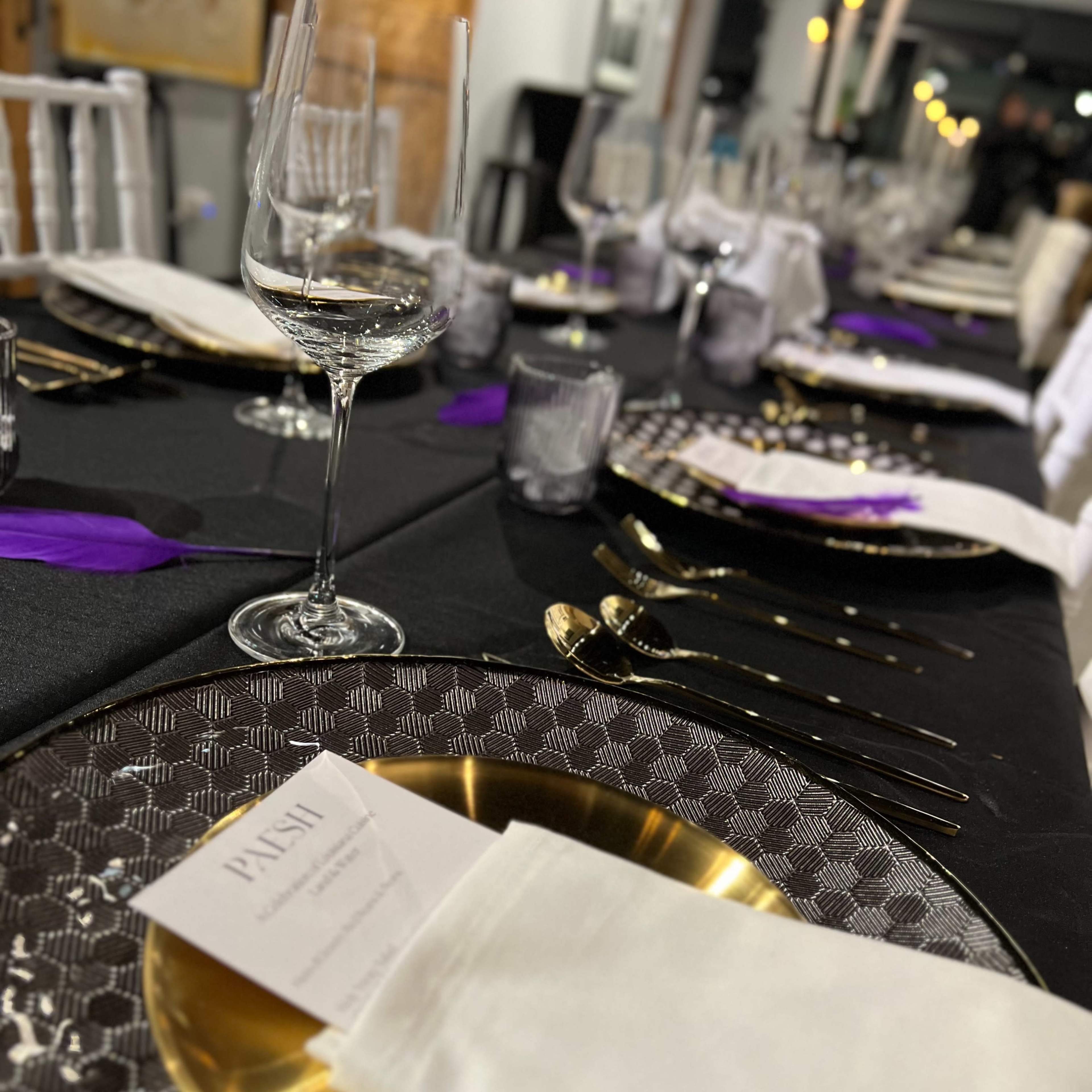 A dining table is set with elegant glassware, golden cutlery, and decorative place settings featuring purple accents.