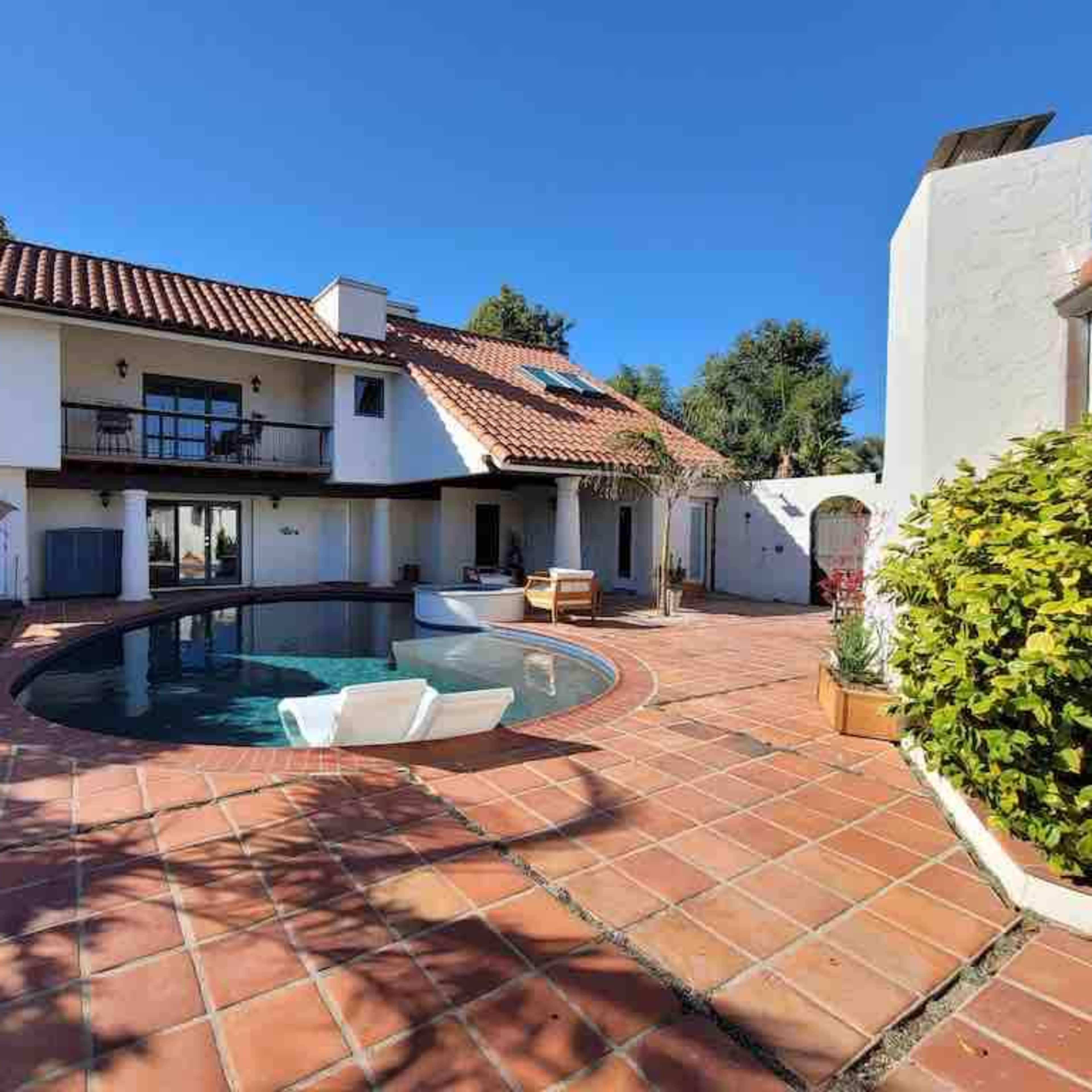 A house with a red-tiled roof surrounds a circular swimming pool, surrounded by a paved patio and landscaped greenery.