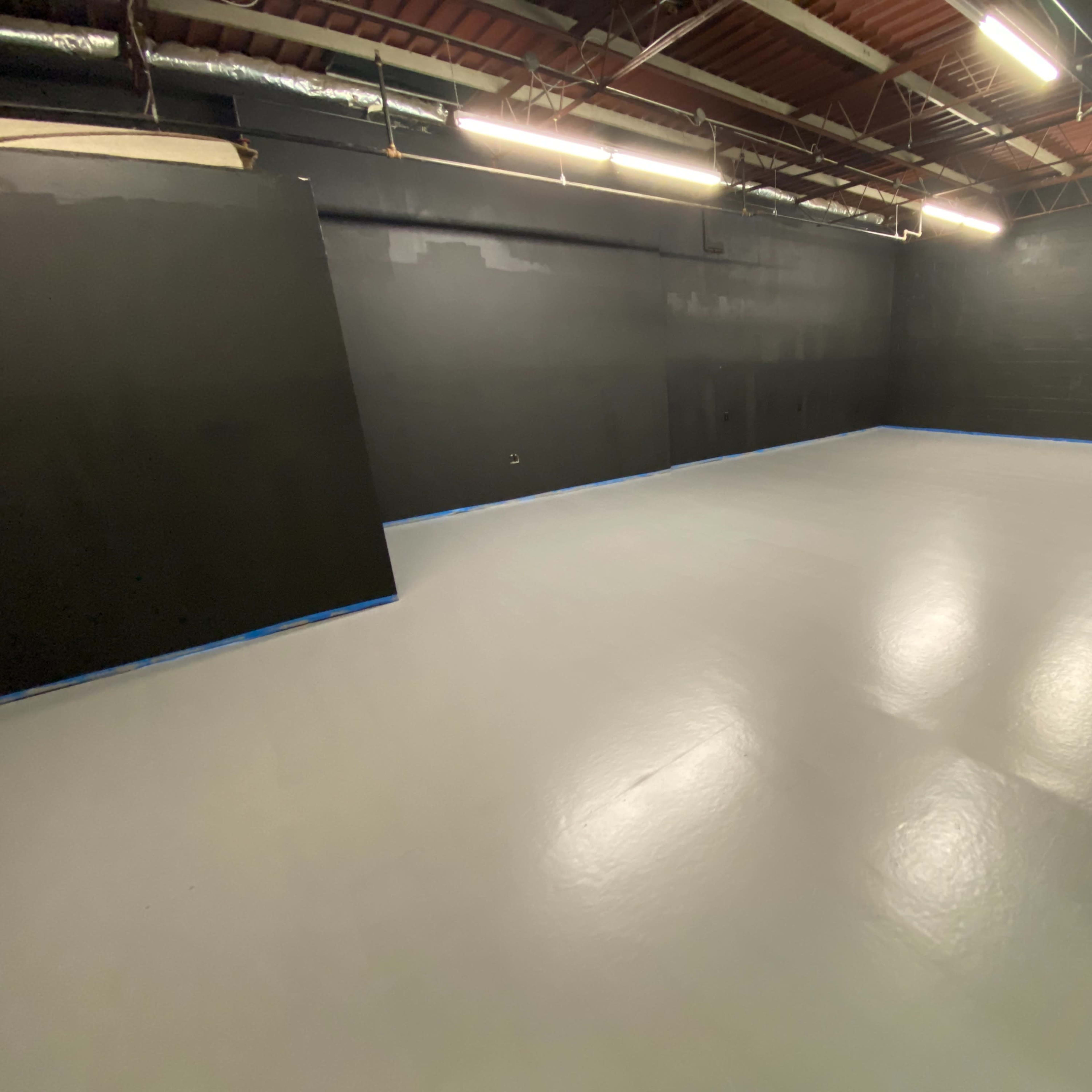 A large, empty room with a gray floor and black walls, illuminated by overhead lights.
