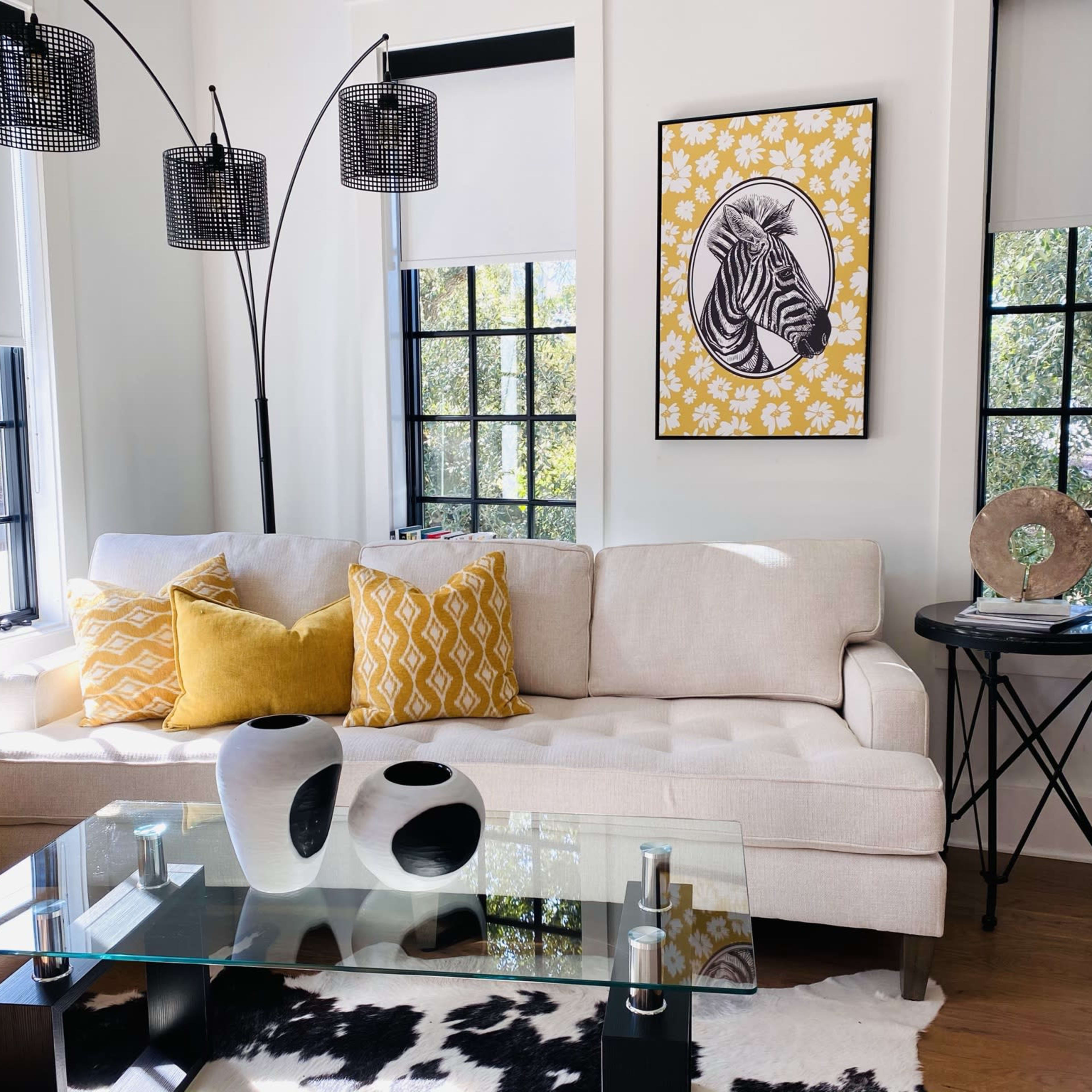 A modern living room featuring a beige sofa with yellow patterned pillows, a glass coffee table, and a zebra print artwork on the wall.