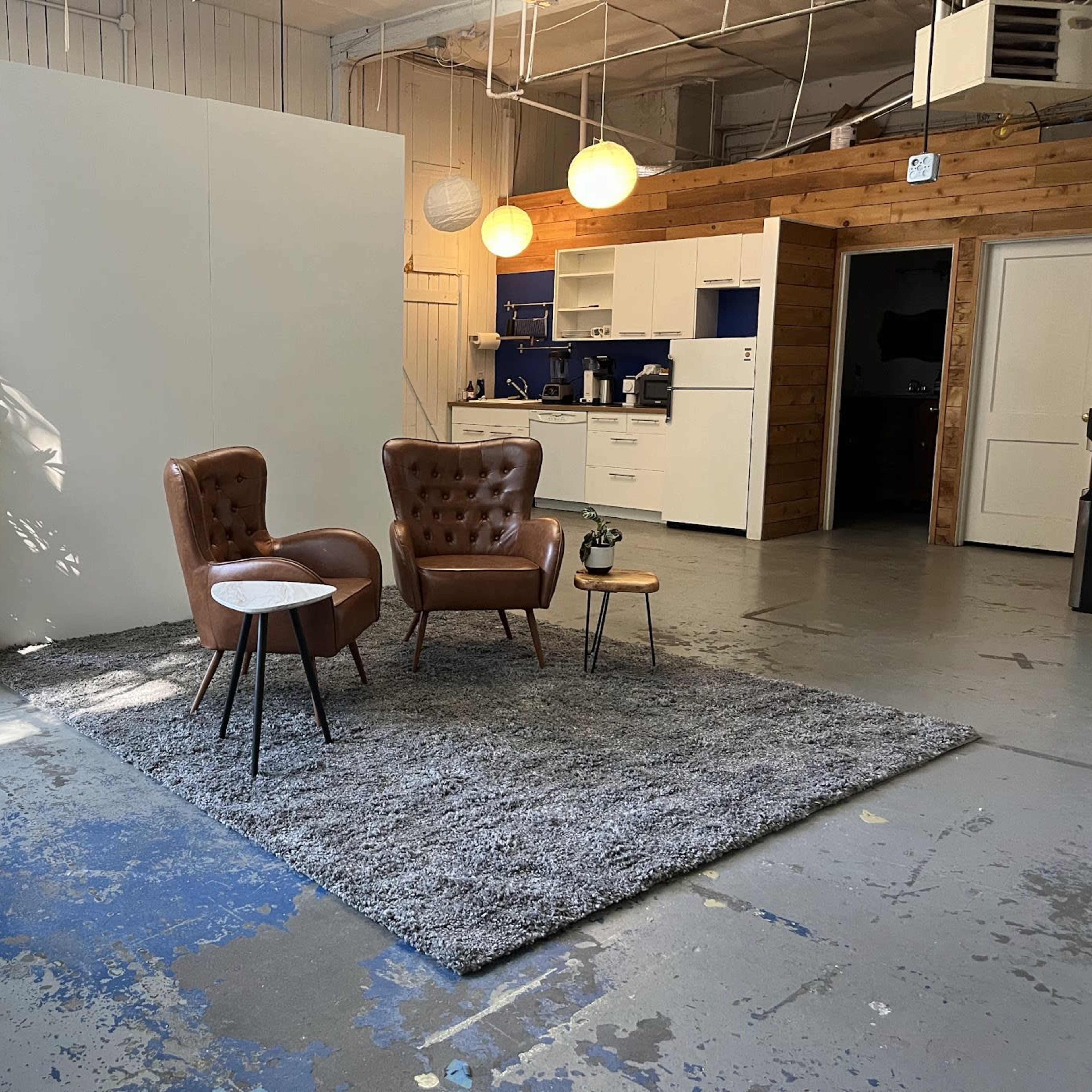 A sparse interior space featuring two brown leather chairs, a small side table, and a large rug, with a white wall in the background and a kitchen area visible to one side.