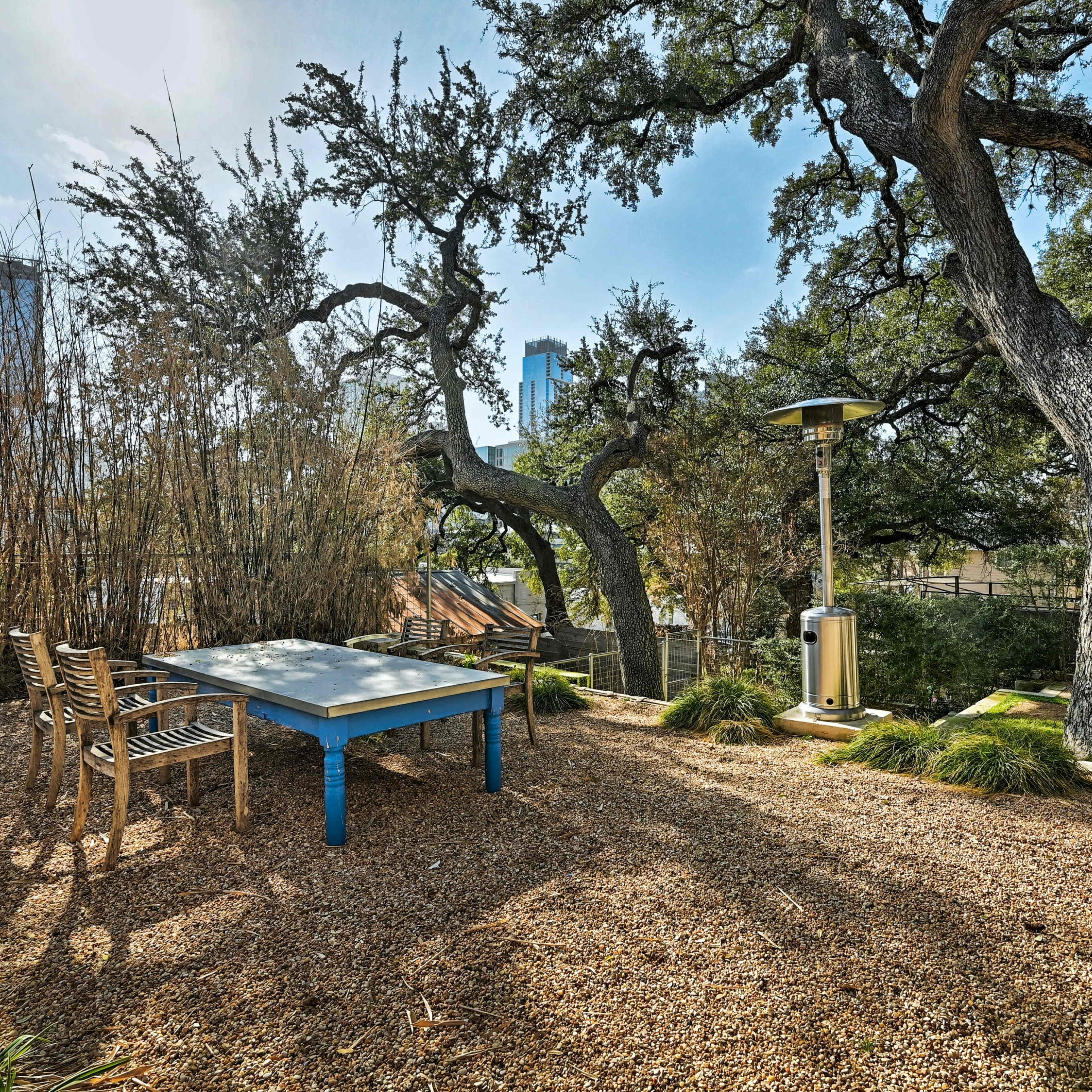 A blue table with wooden chairs sits in a gravel area surrounded by trees and tall buildings in the background.