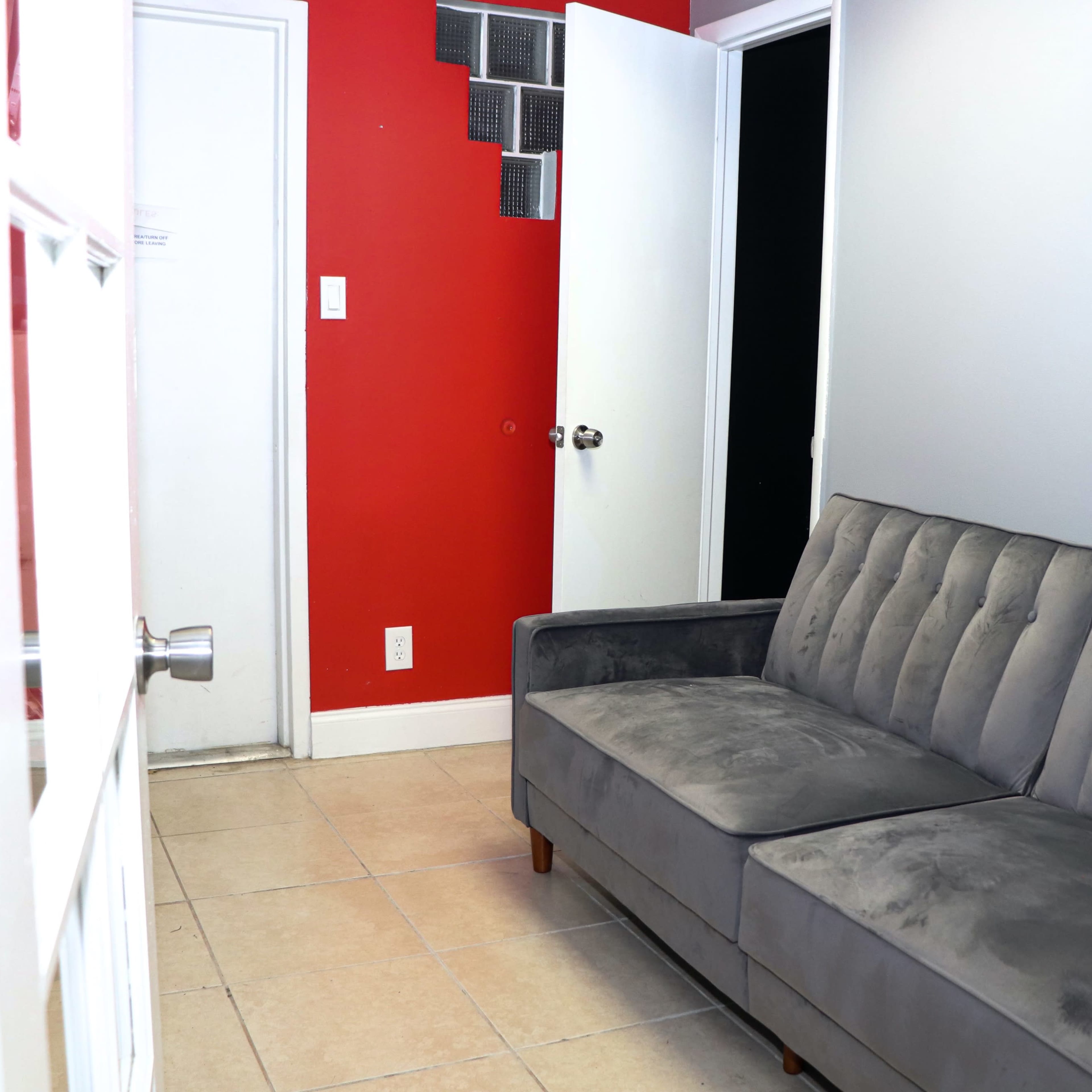A gray tufted sofa is positioned against a wall in a small room with red and white walls, featuring two doors and a tiled floor.