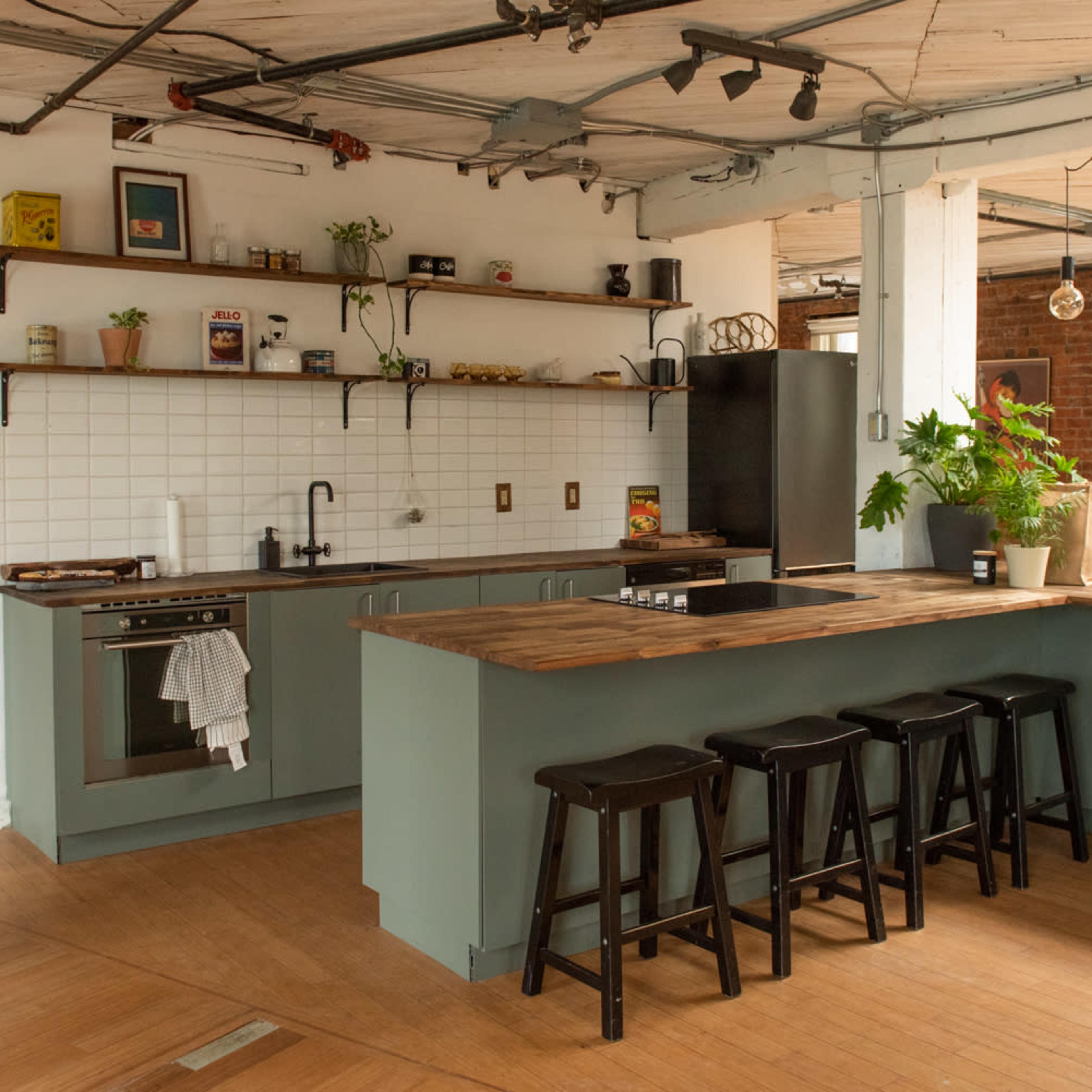 A modern kitchen features a wooden bar counter with four black stools, open shelving, and a mix of plants along the walls.