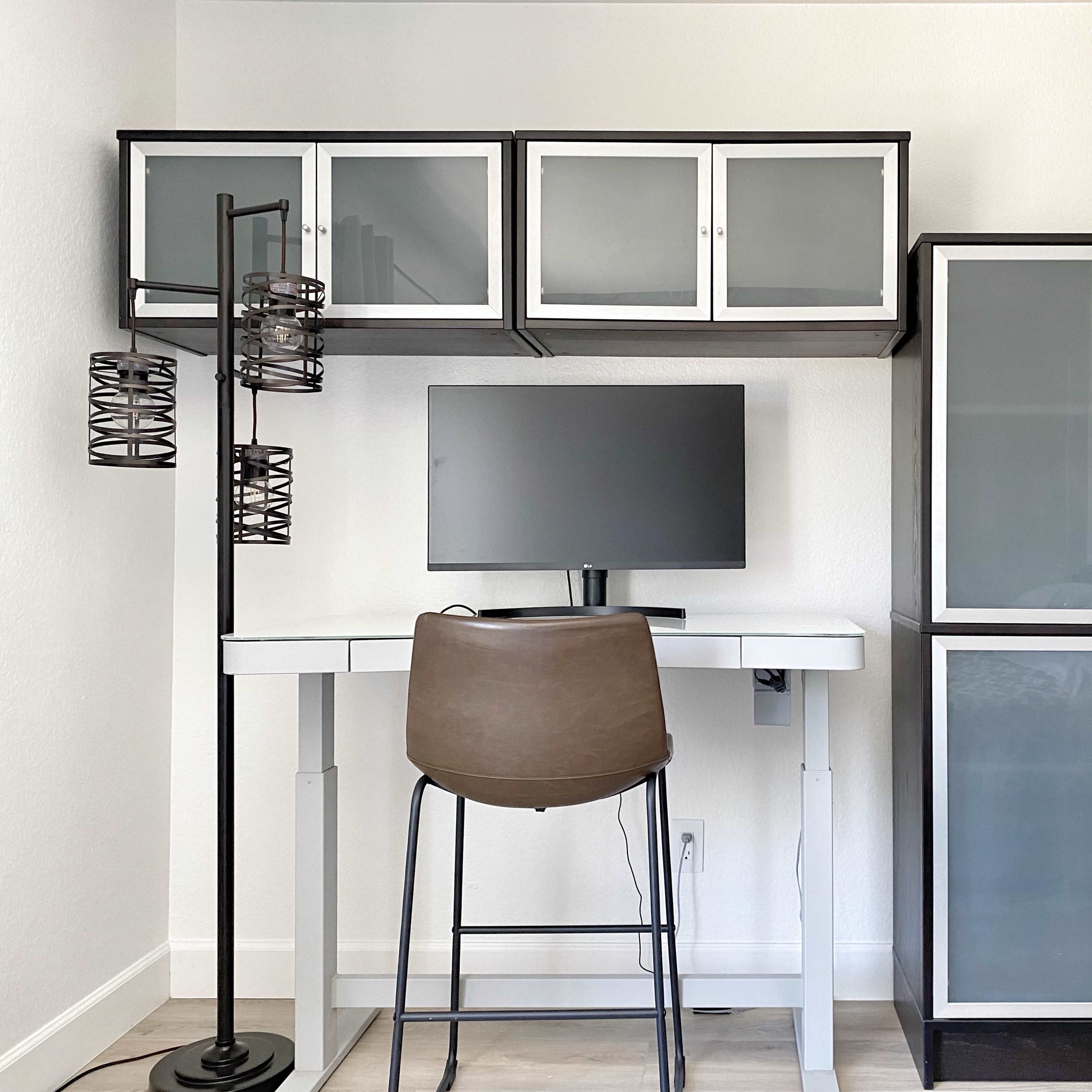 A minimalistic home office setup with a desk, a computer monitor, a chair, and cabinets on the wall and beside the desk.