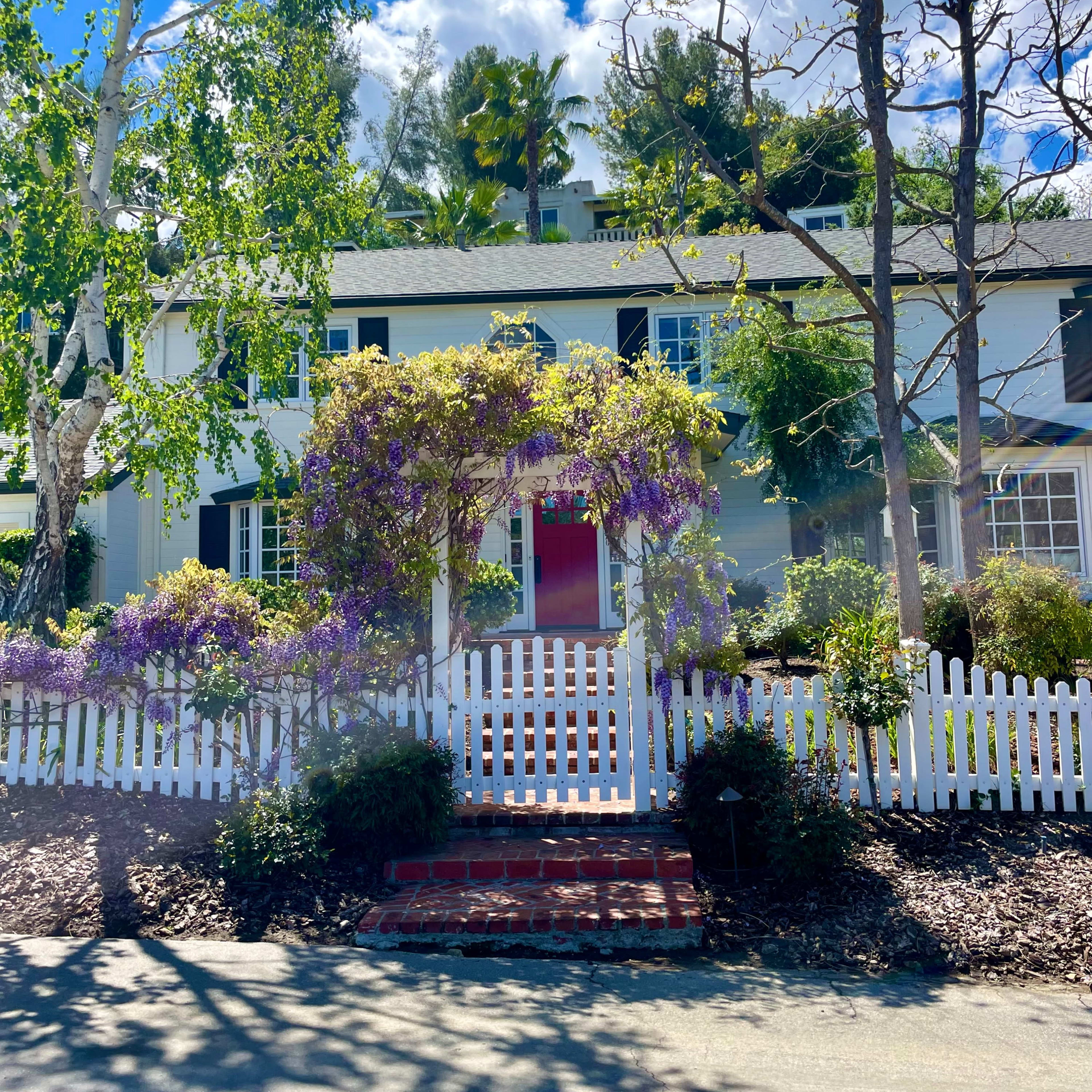 A two-story white house with a red door is surrounded by lush greenery and a white picket fence, with purple flowering vines and a paved walkway leading to the entrance.