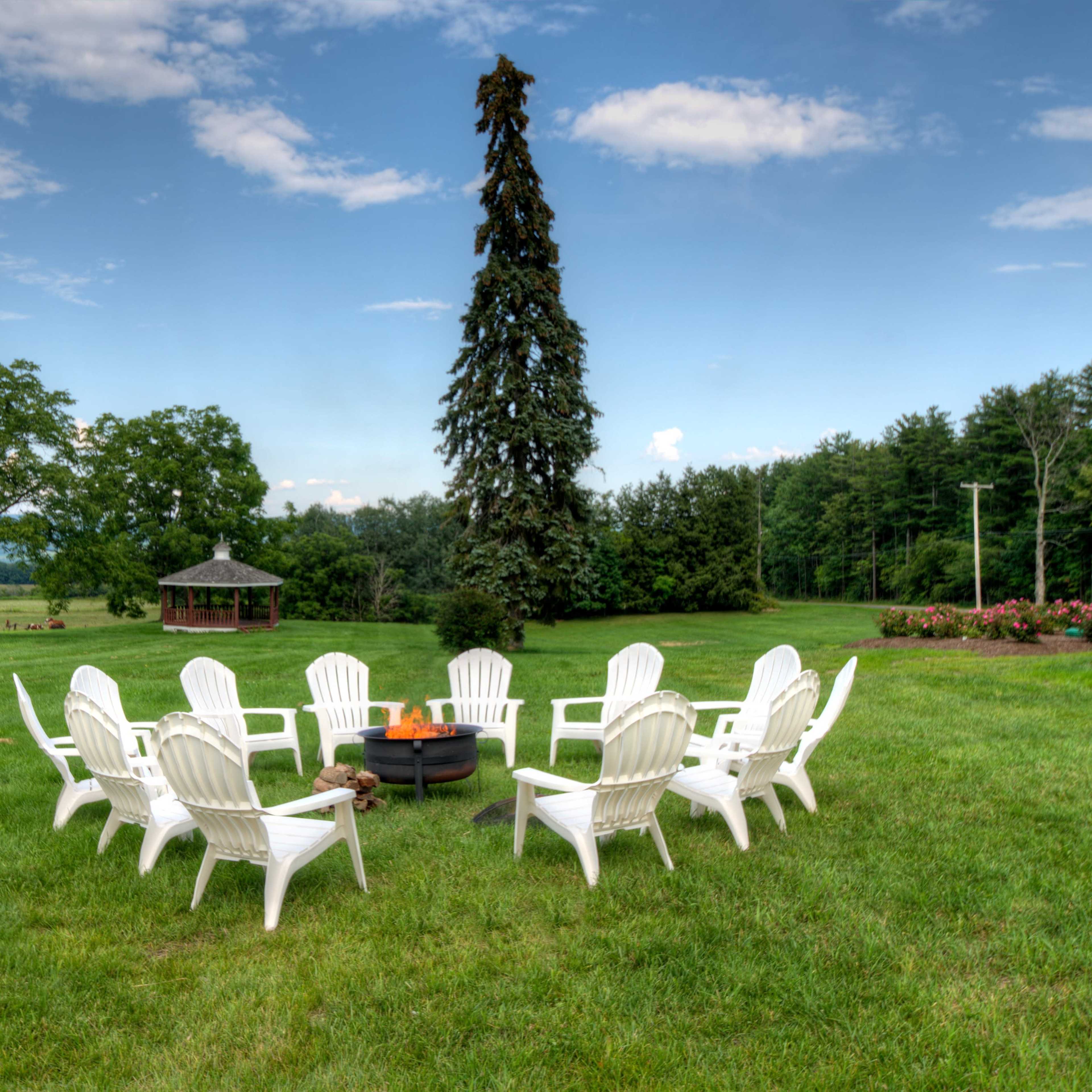 A circle of white adirondack chairs surrounds a fire pit on a grassy area, with a gazebo and trees in the background.