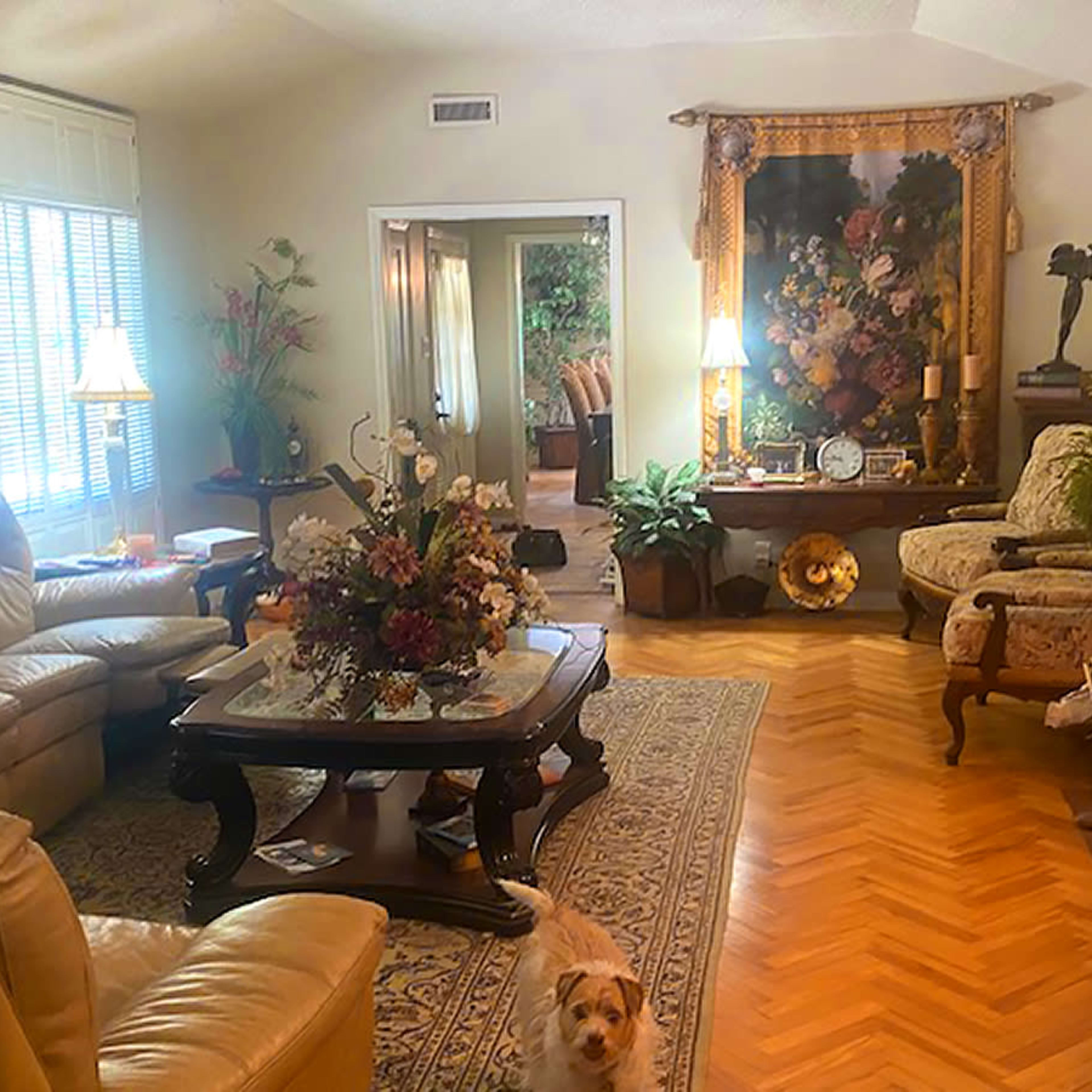 The living room features a mix of leather and upholstered furniture, a floral arrangement on a wooden table, and a patterned rug on a herringbone floor.