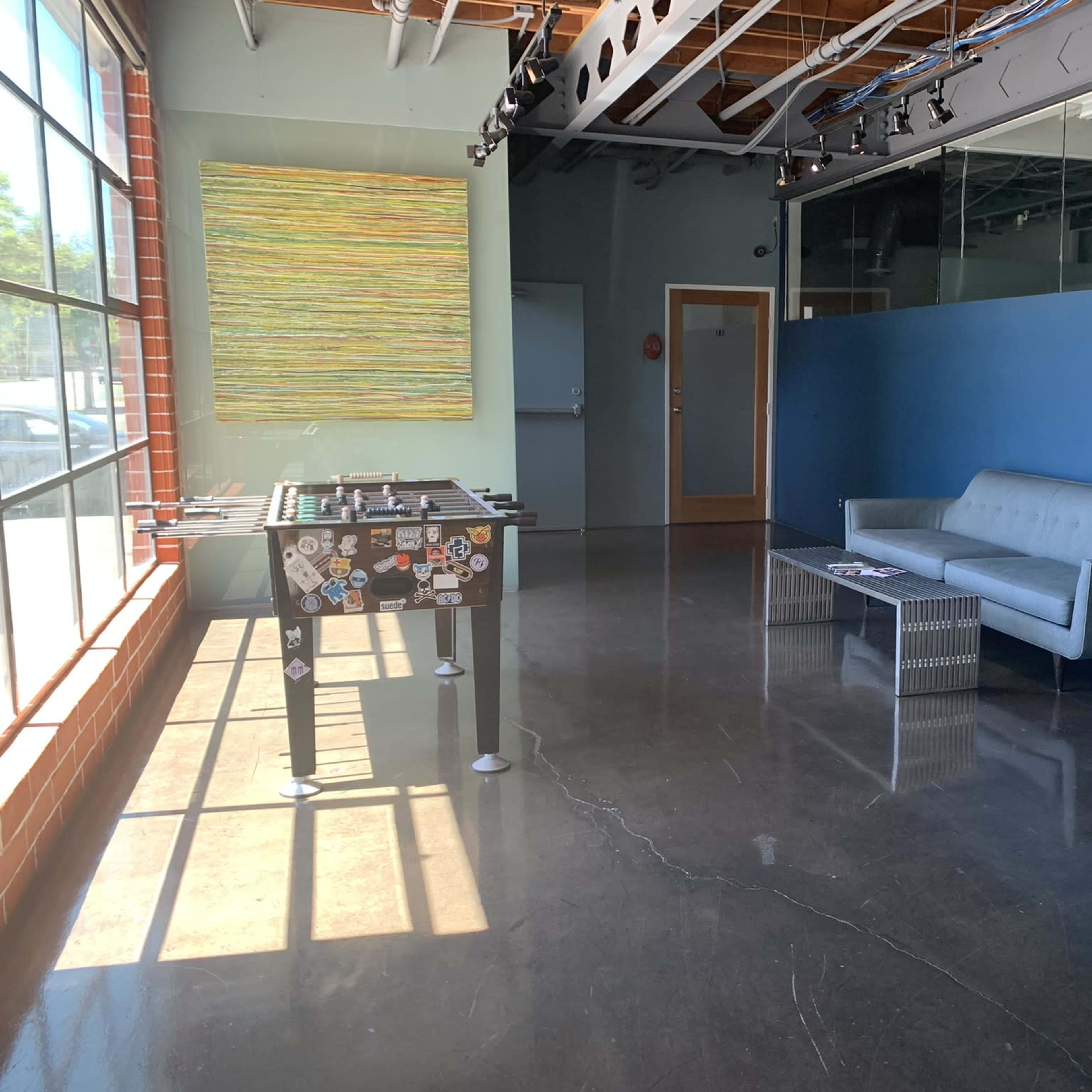 An empty room with a modern gray couch, a foosball table decorated with stickers, and large windows allowing natural light to enter.