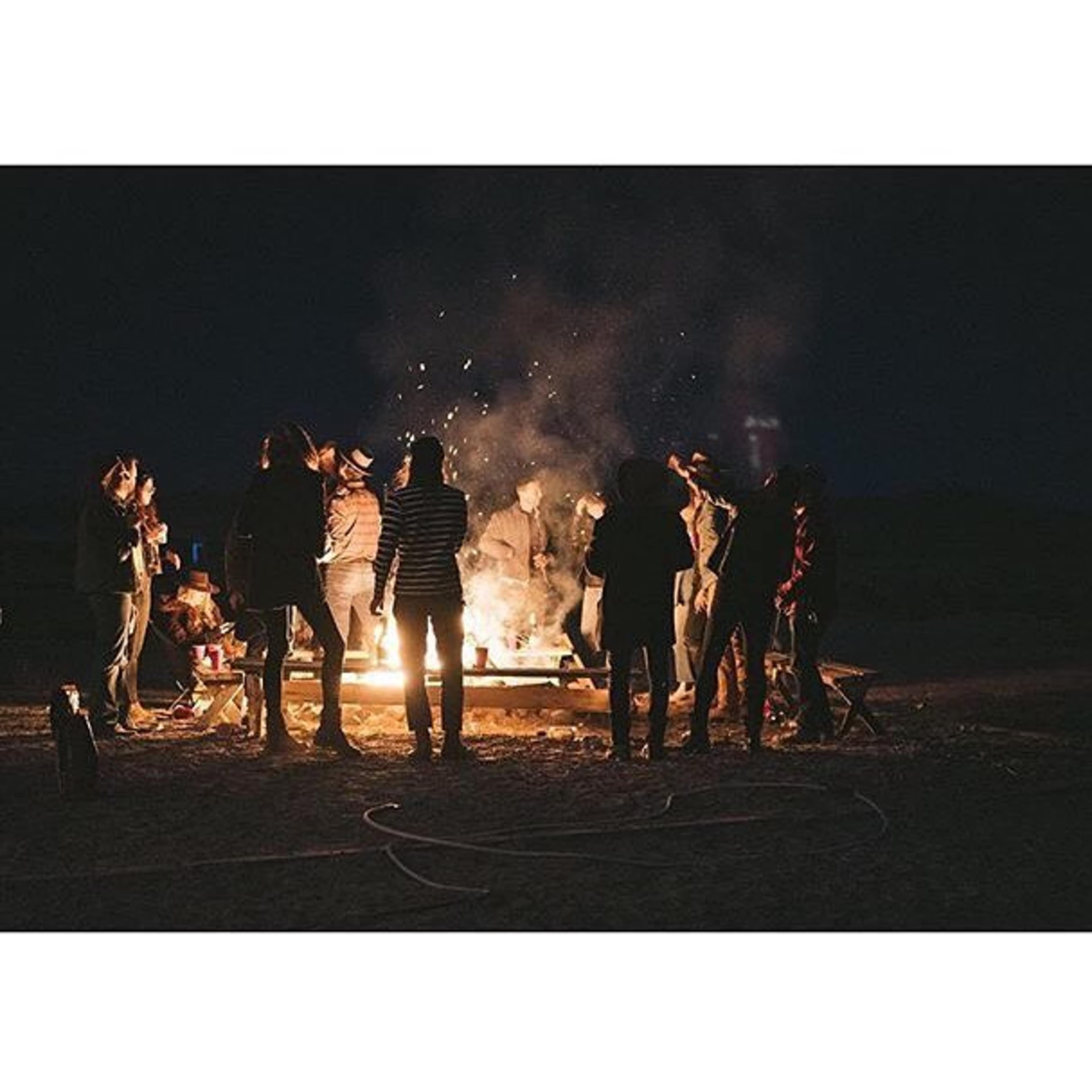 A group of people gathers around a large campfire at night, with sparks rising into the dark sky.