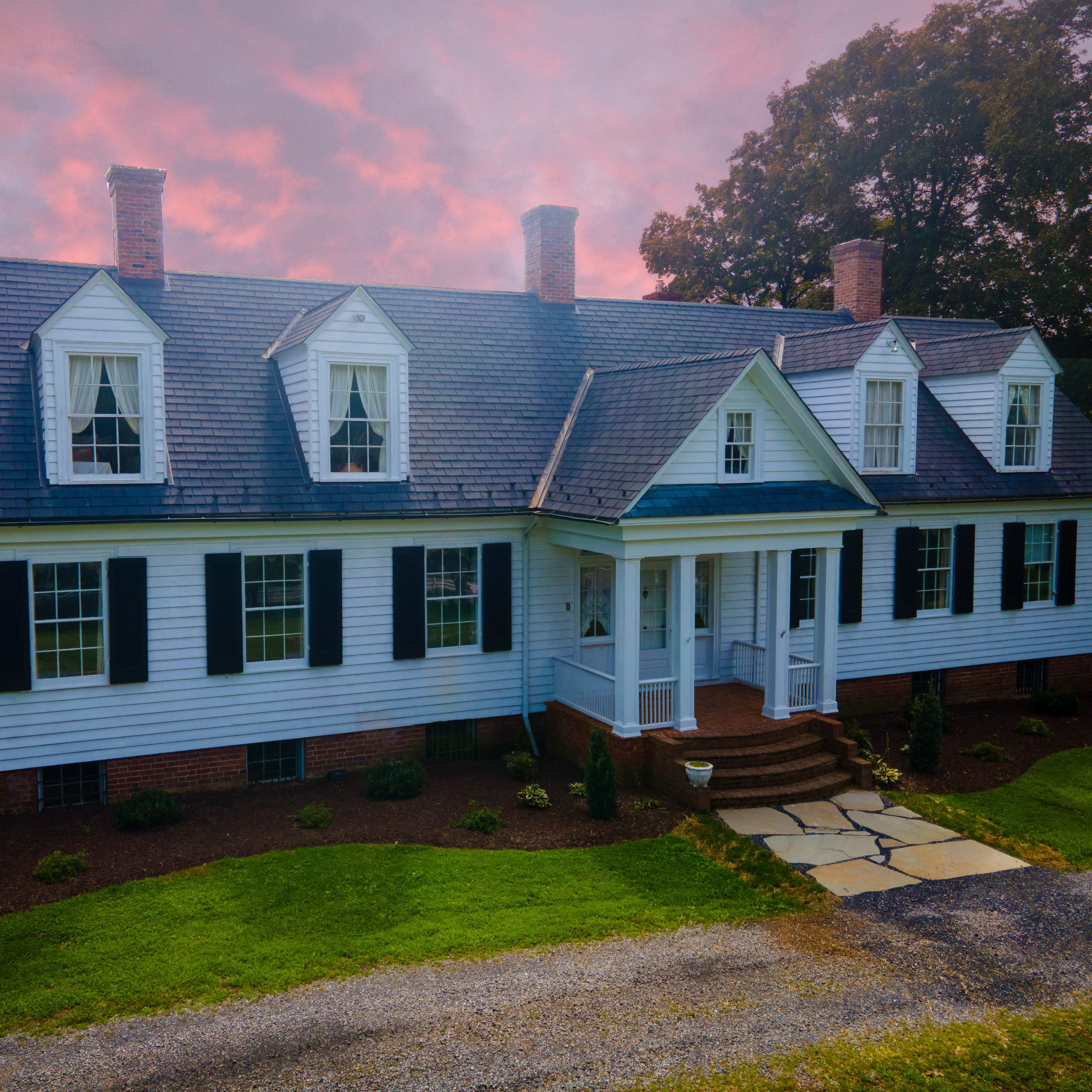 A two-story white colonial-style house features a symmetrical facade, with dormer windows and a front porch, set against a backdrop of a colorful sky.
