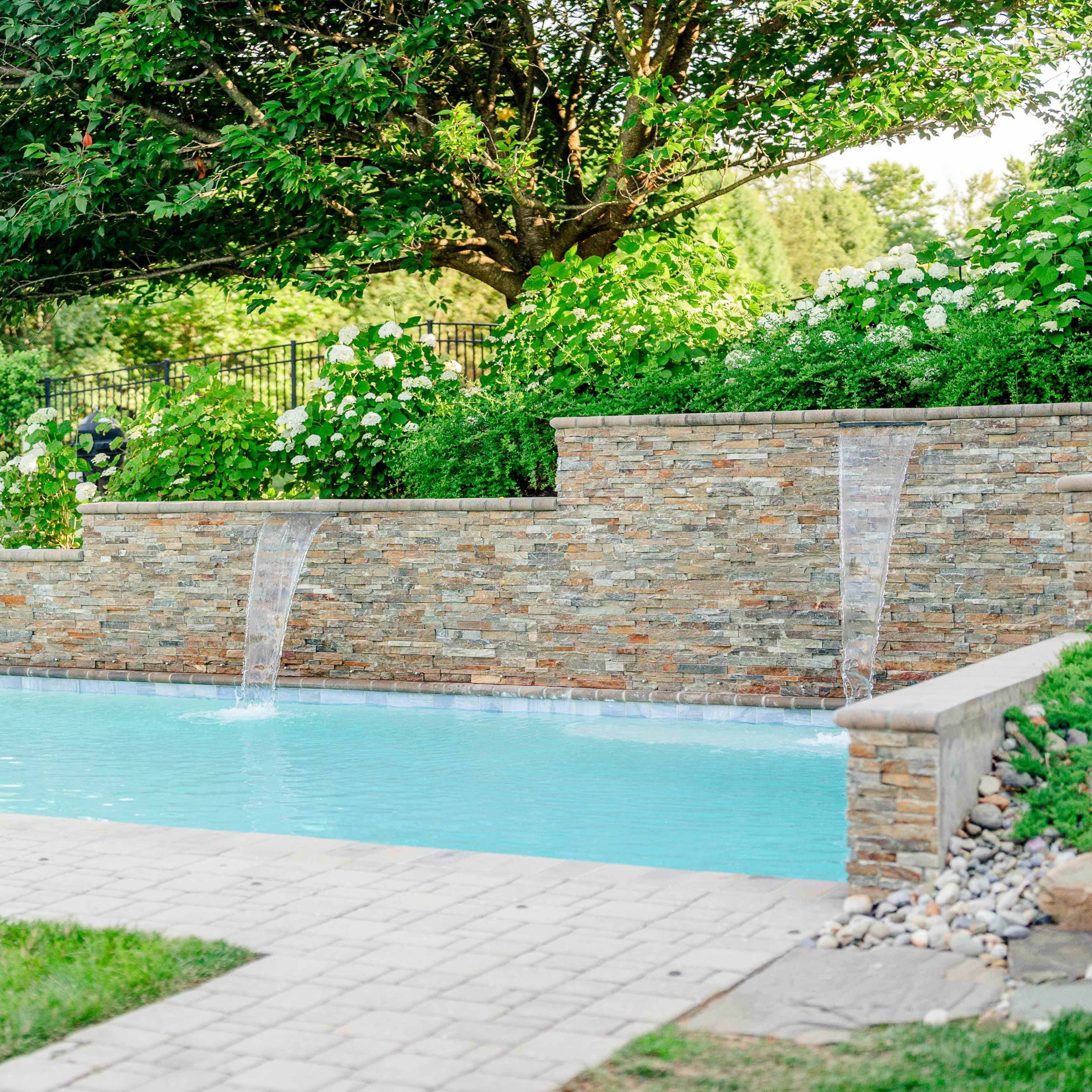 A backyard swimming pool with two water features flowing into it, surrounded by greenery and stone walls.