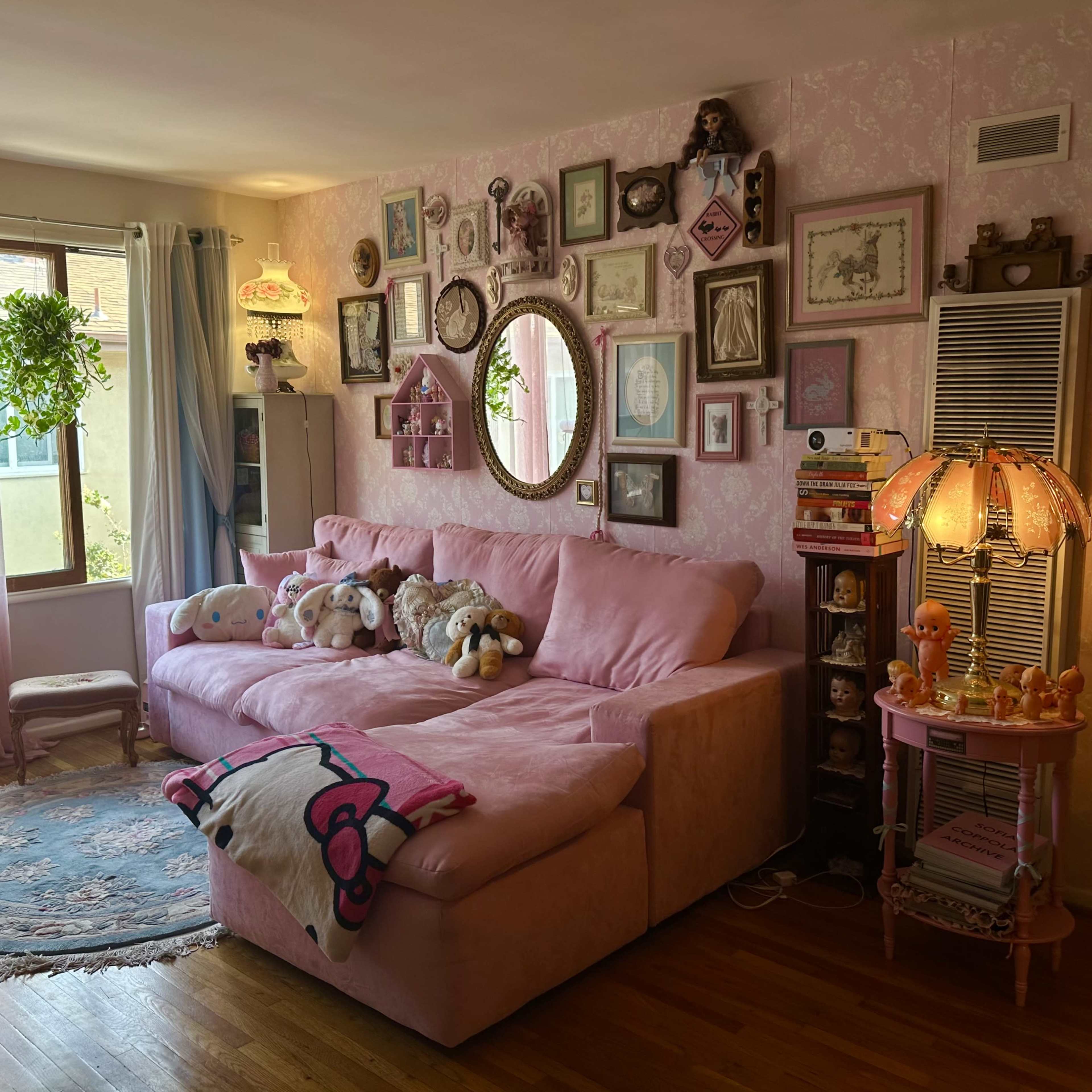 A cozy living room with a pink sectional sofa surrounded by framed artwork and decorative objects, including plants and a lamp.