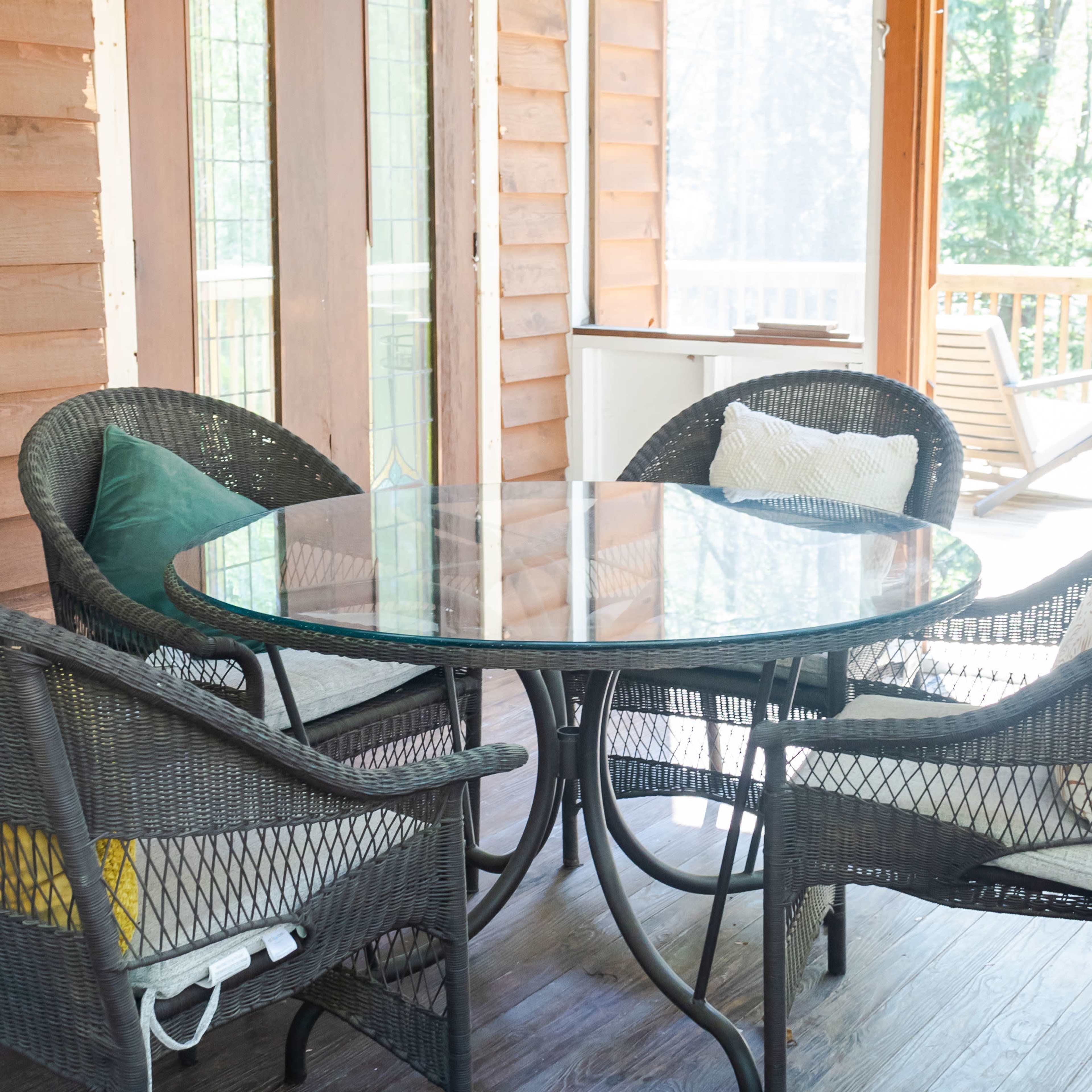 A round glass table is set with four wicker chairs on a porch with wooden walls and large windows overlooking a wooded area.