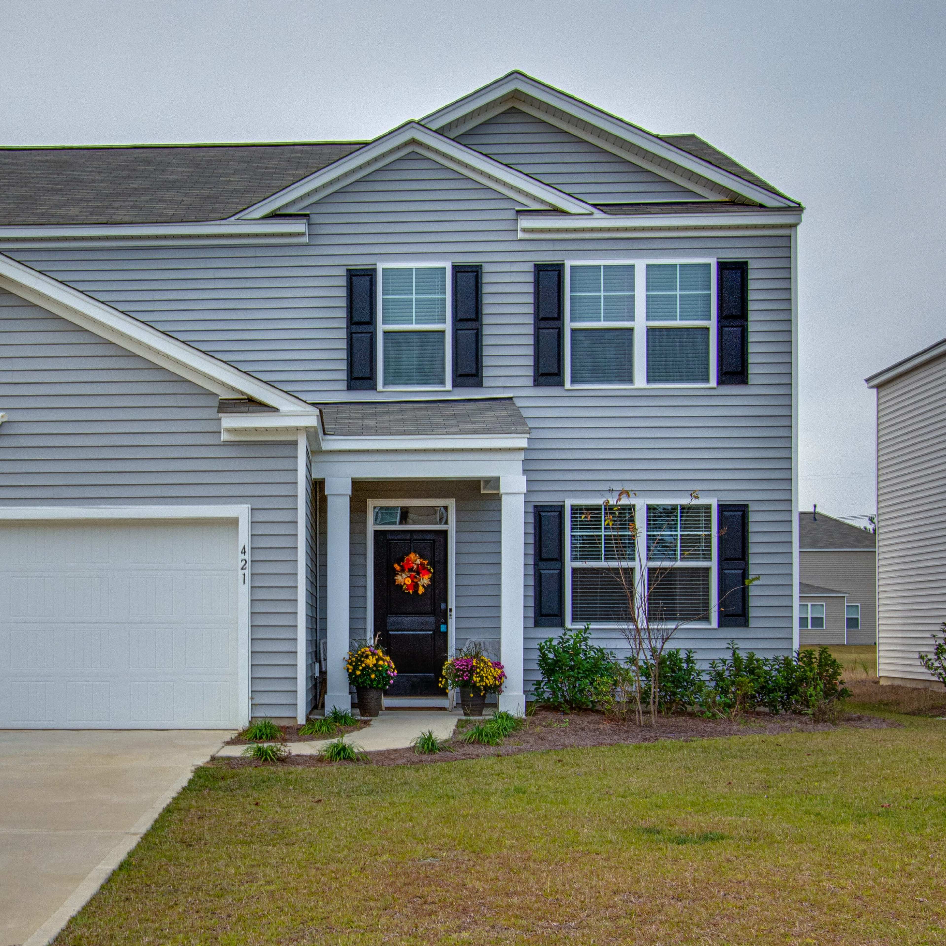 A two-story gray house with black shutters and a white garage door features a front porch adorned with a wreath and flower beds on either side.