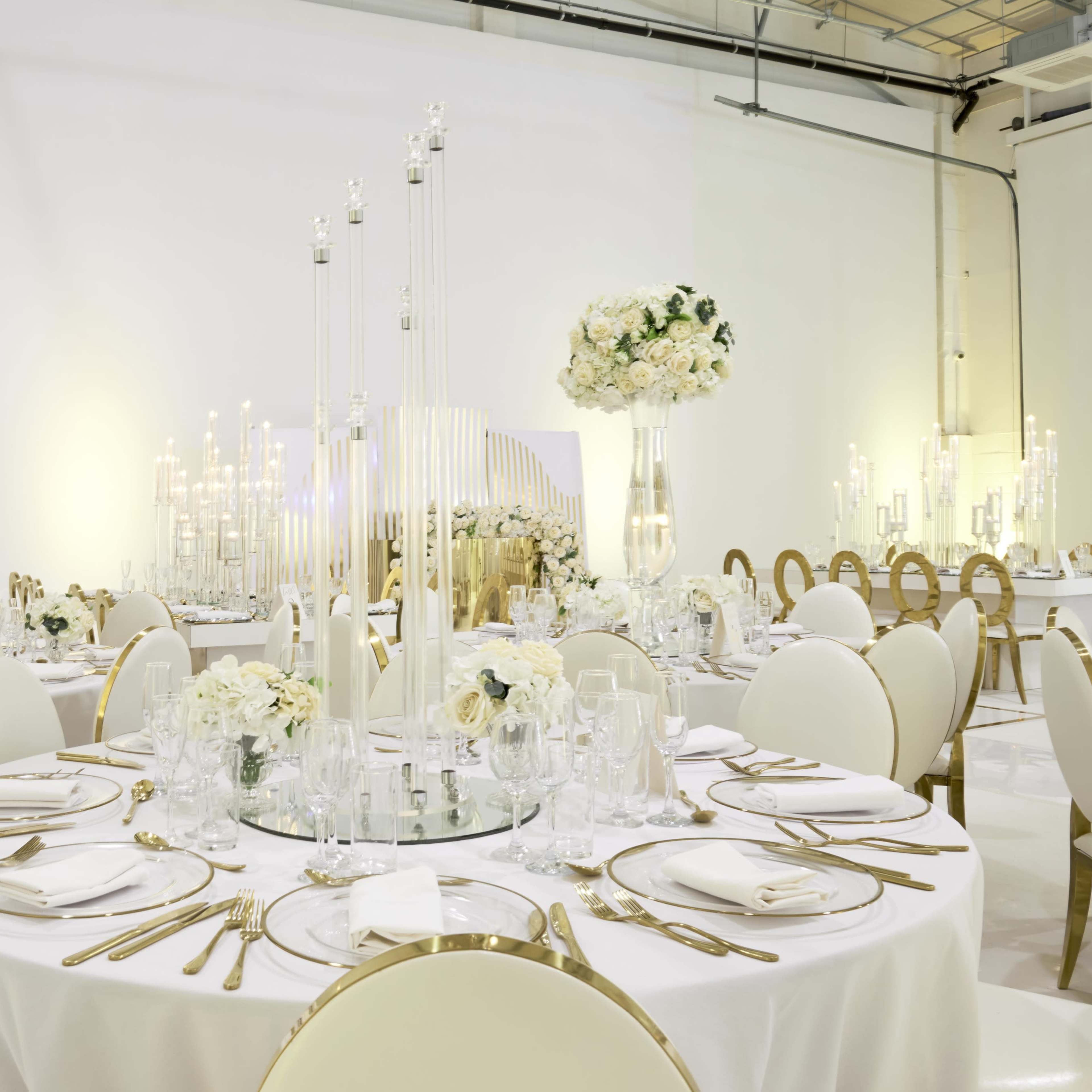 A banquet hall is set with elegantly arranged round tables adorned with white tablecloths, gold cutlery, and floral centerpieces.