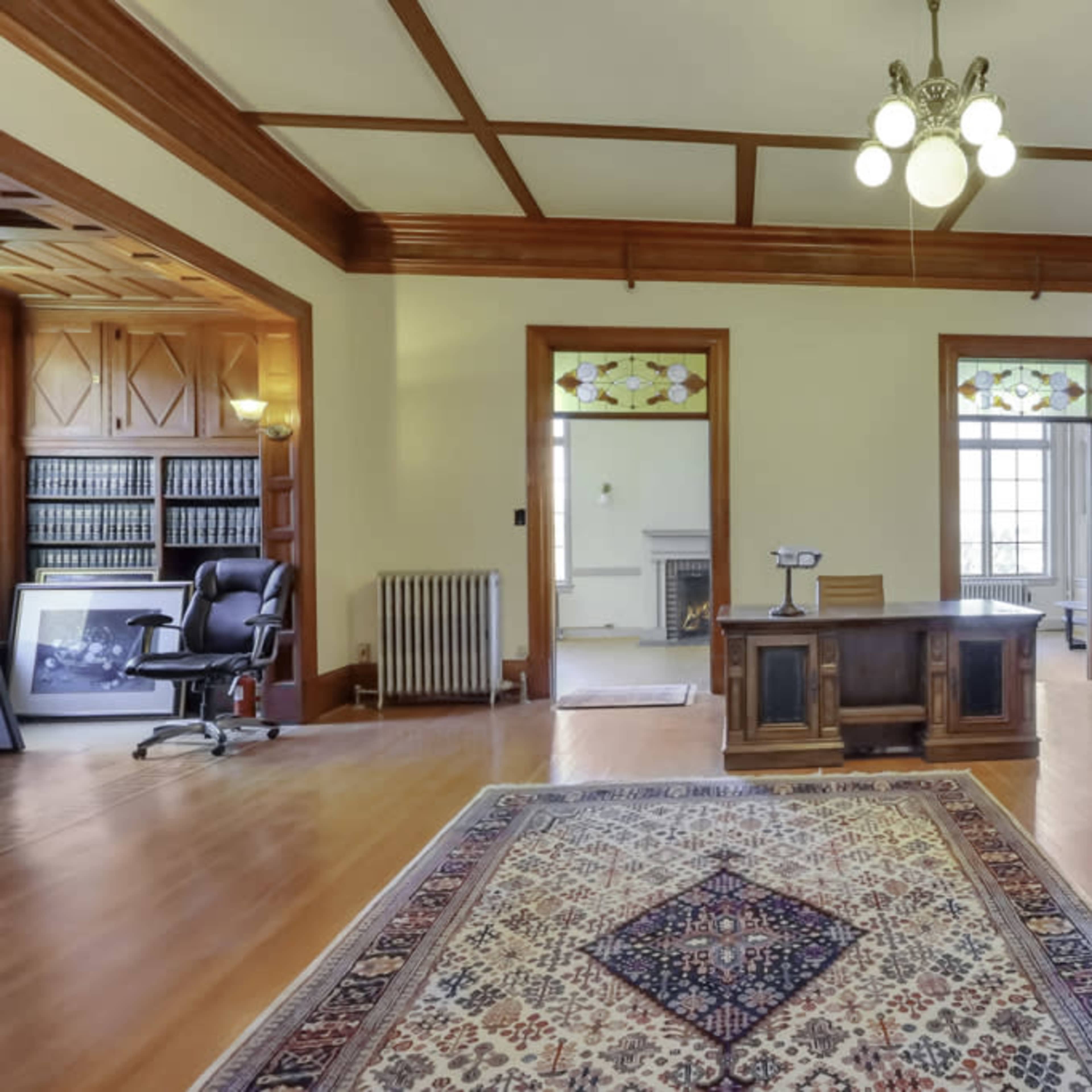 A spacious office with a wooden desk, a black office chair, bookshelves, and large windows allowing natural light to enter.