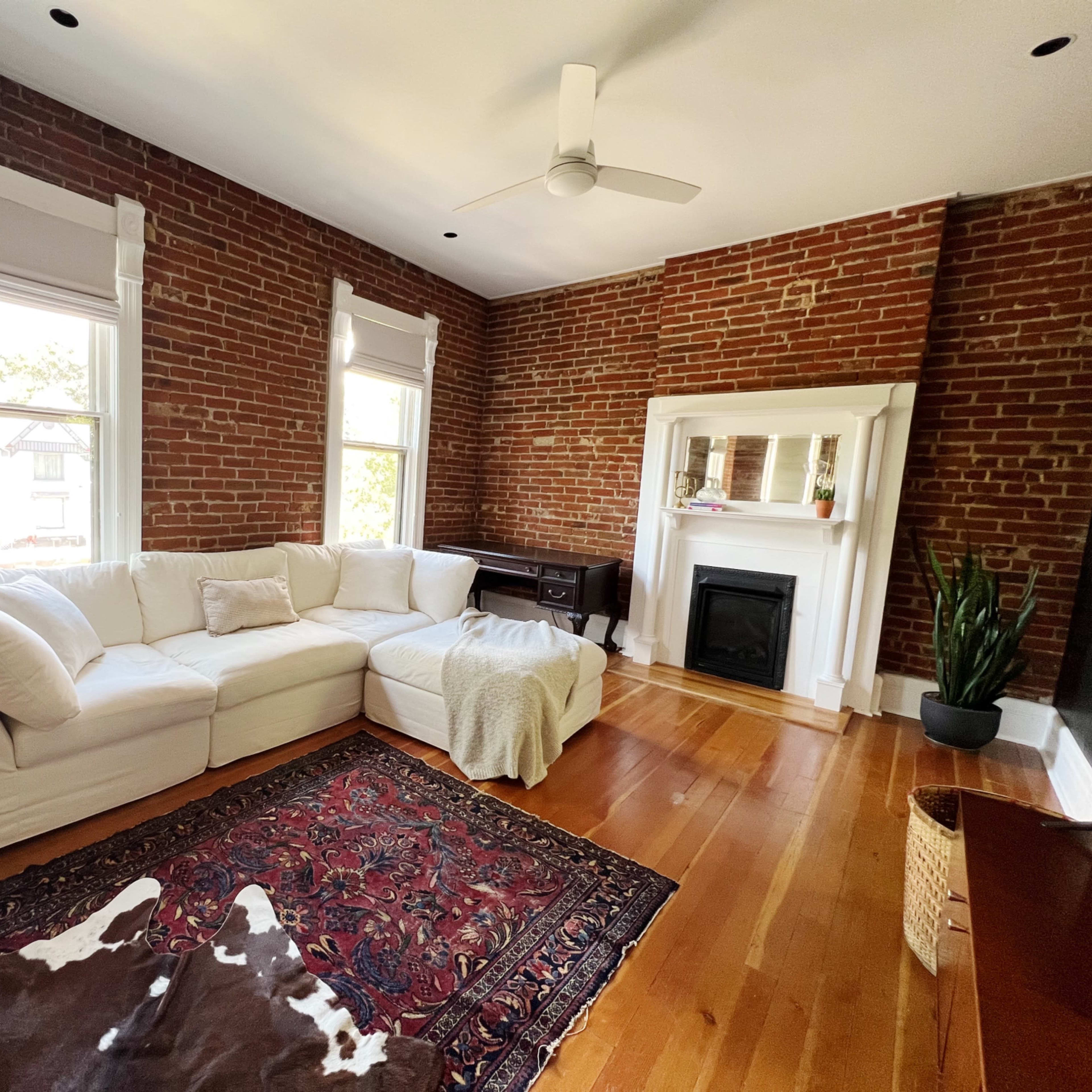 The living room features a white sectional sofa, a fireplace, and exposed brick walls, with hardwood flooring and a patterned rug.