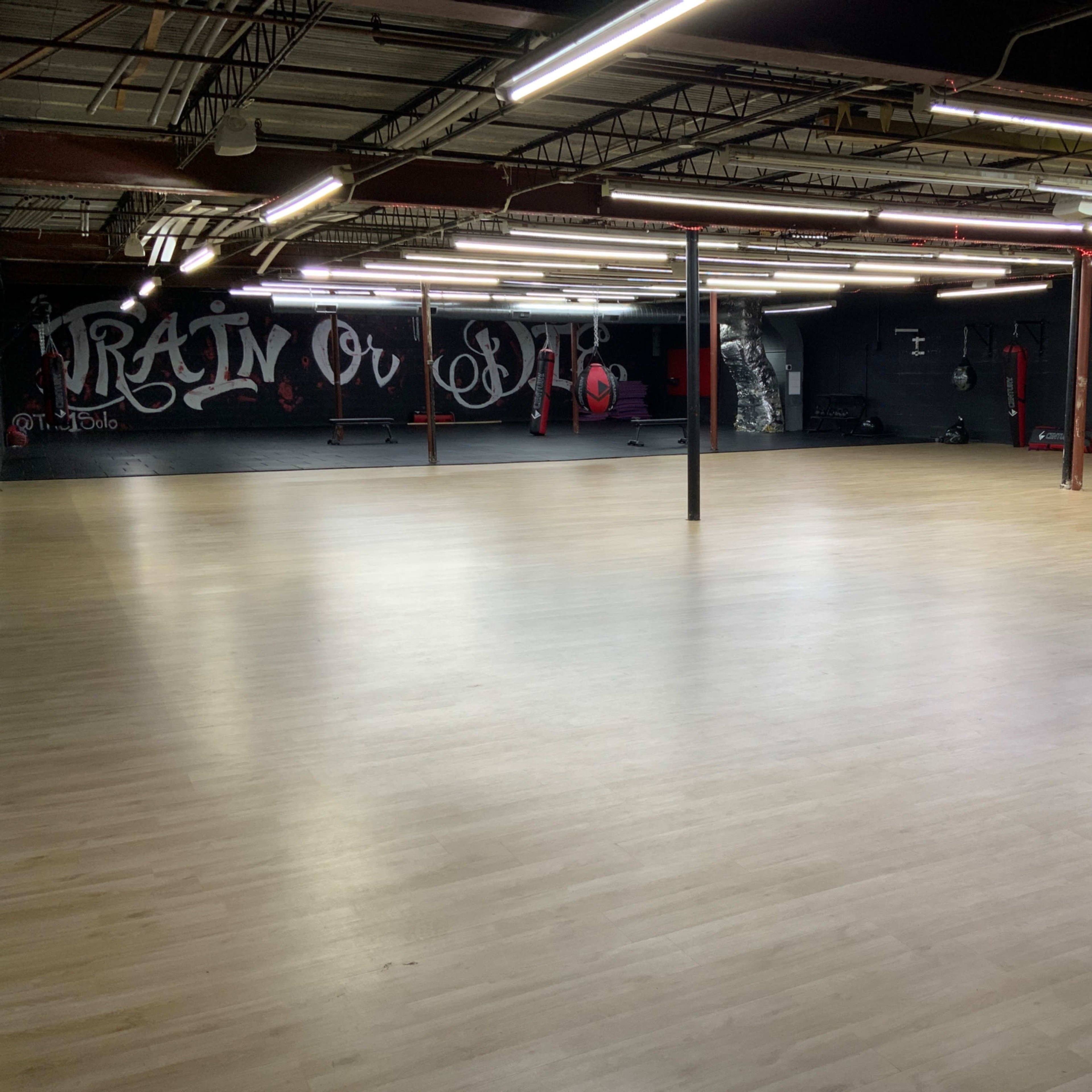 A large, empty gym space with smooth wooden flooring and a graffiti-style mural on the wall.