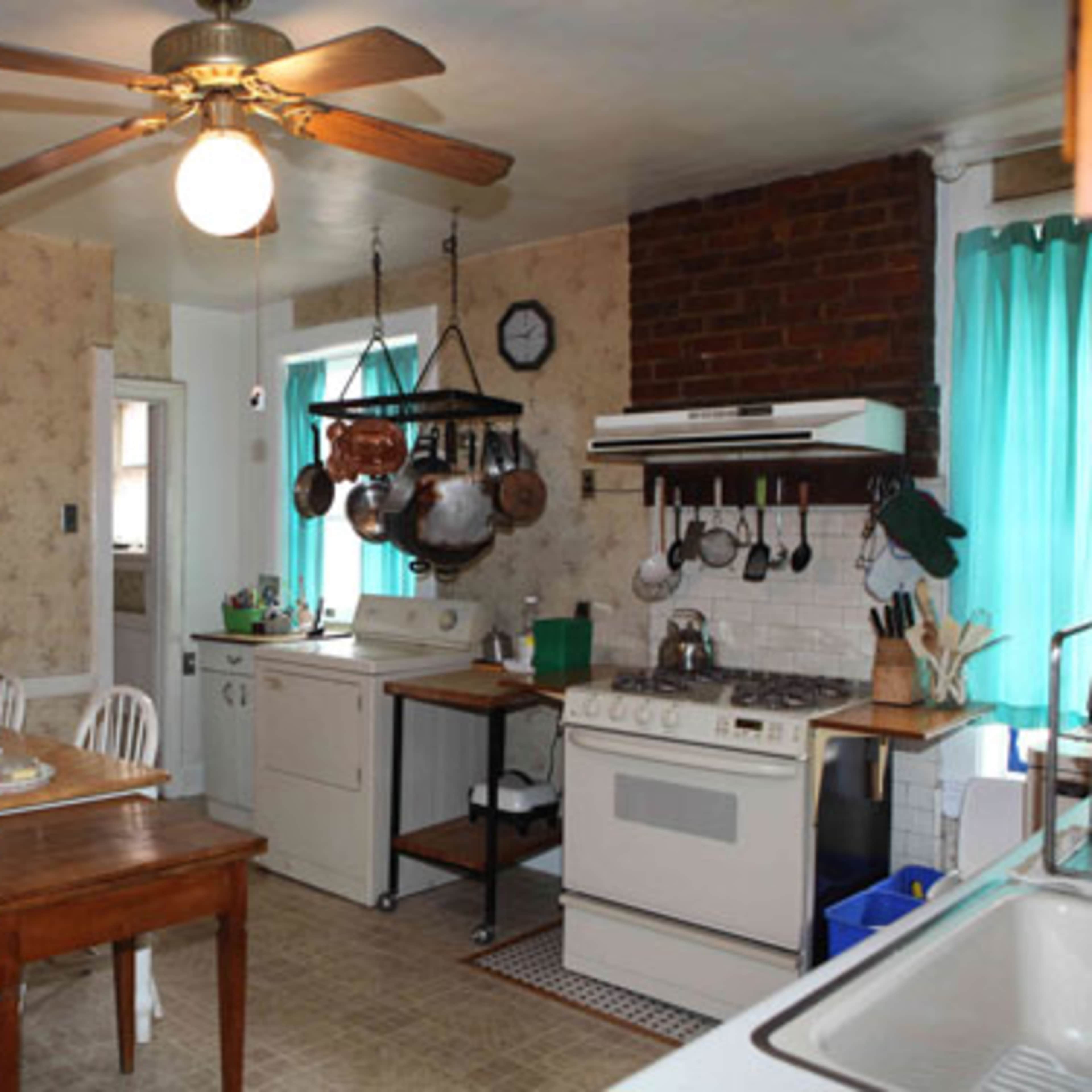 A modest kitchen with a wooden table and chairs, a stove, a refrigerator, and various pots hanging from a rack.