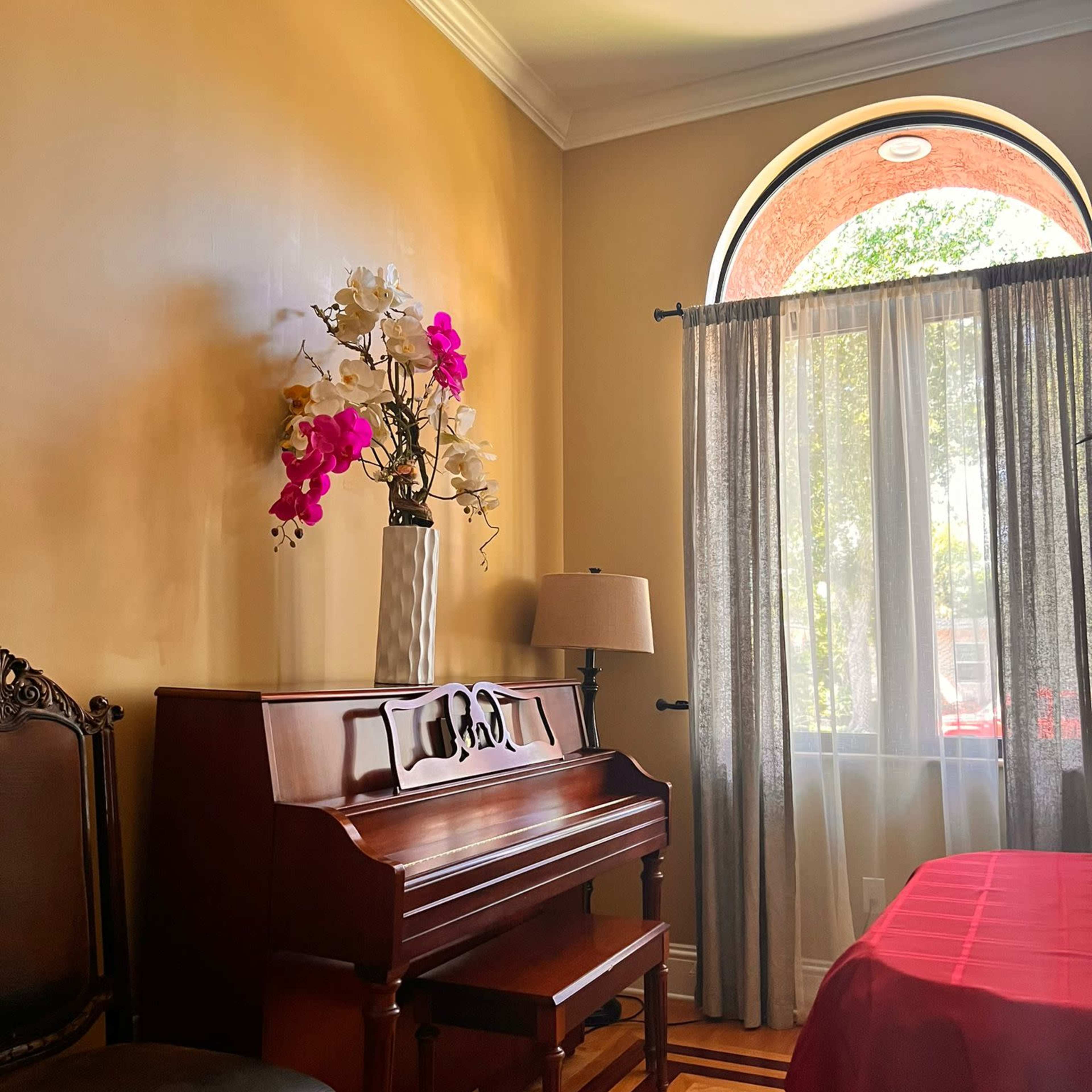 A wooden piano is positioned next to a window adorned with sheer curtains, with a vase of flowers on top and a lamp nearby.